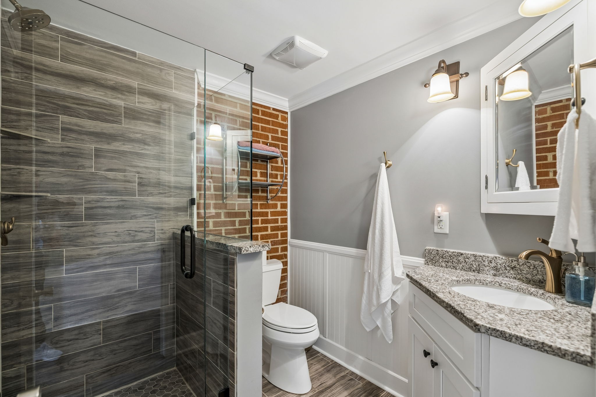 Bathroom with a gray-tiled glass shower, exposed brick accent wall, and granite vanity.