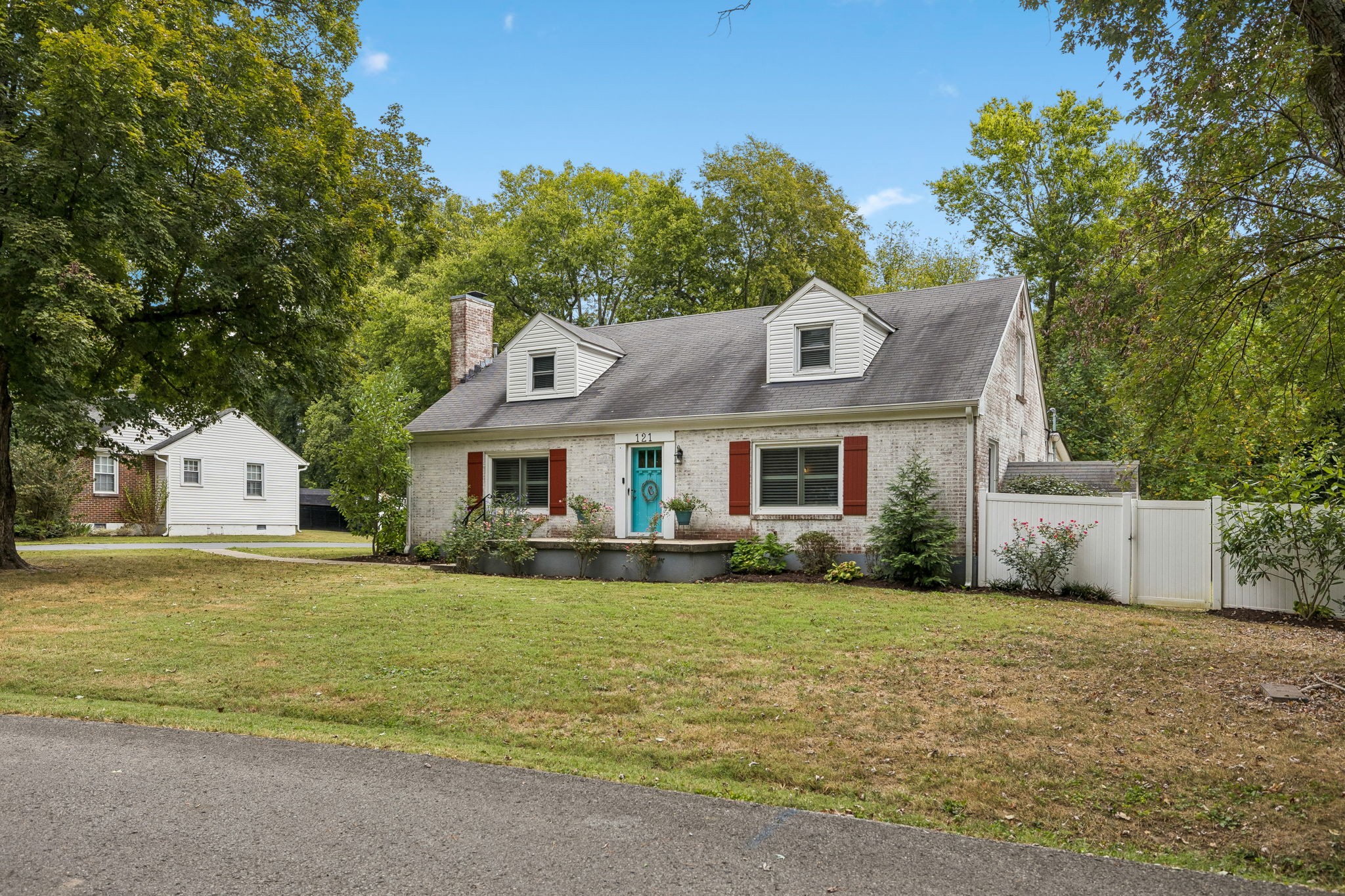 White-brick house with red shutters and a turquoise front door, set among mature trees.