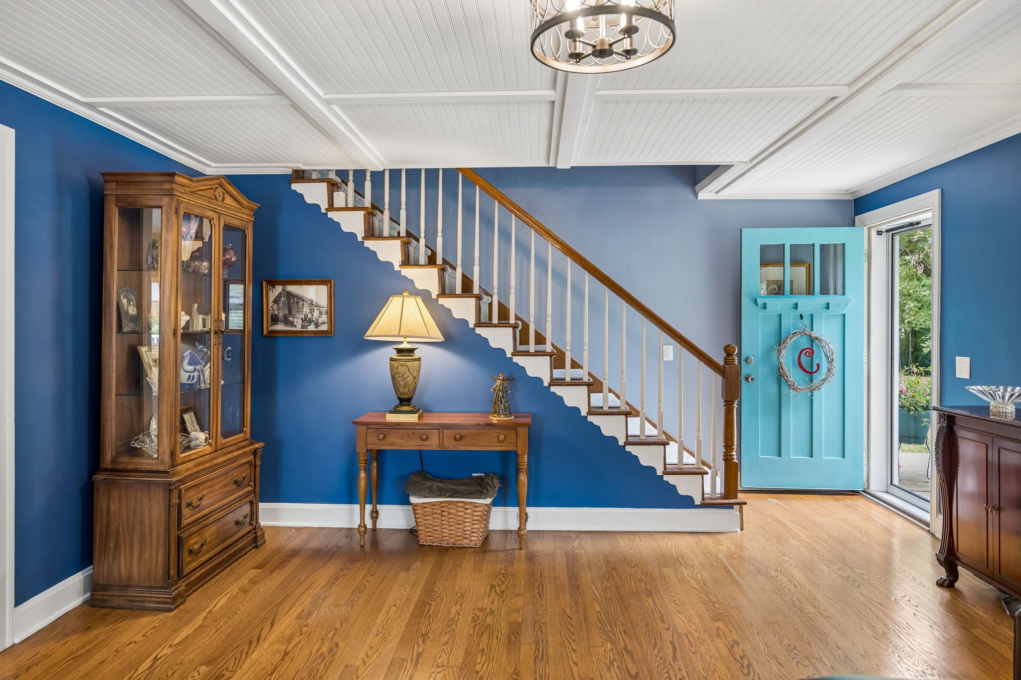 Entry foyer with blue walls, hardwood floors, a diagonal staircase, and an open turquoise front door.