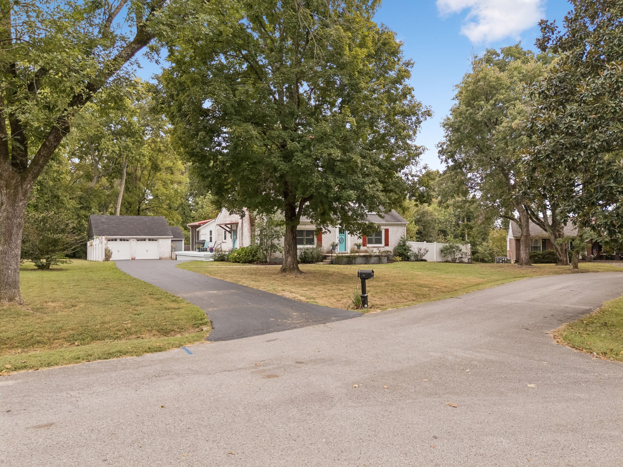 White brick house with a turquoise door and red shutters beneath mature trees, with a detached garage and driveway.