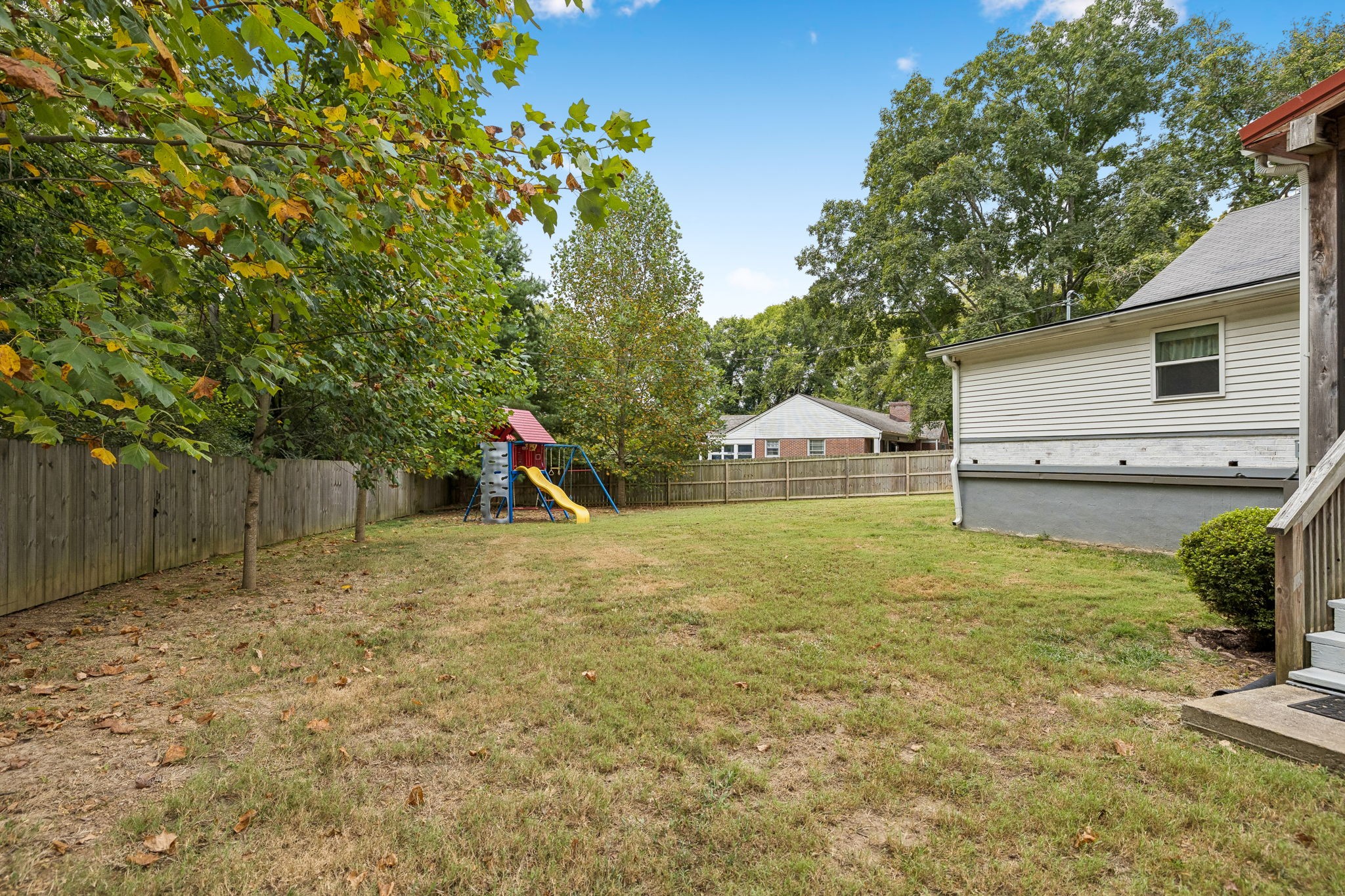 Fenced backyard with a children's playset and slide beneath mature trees.