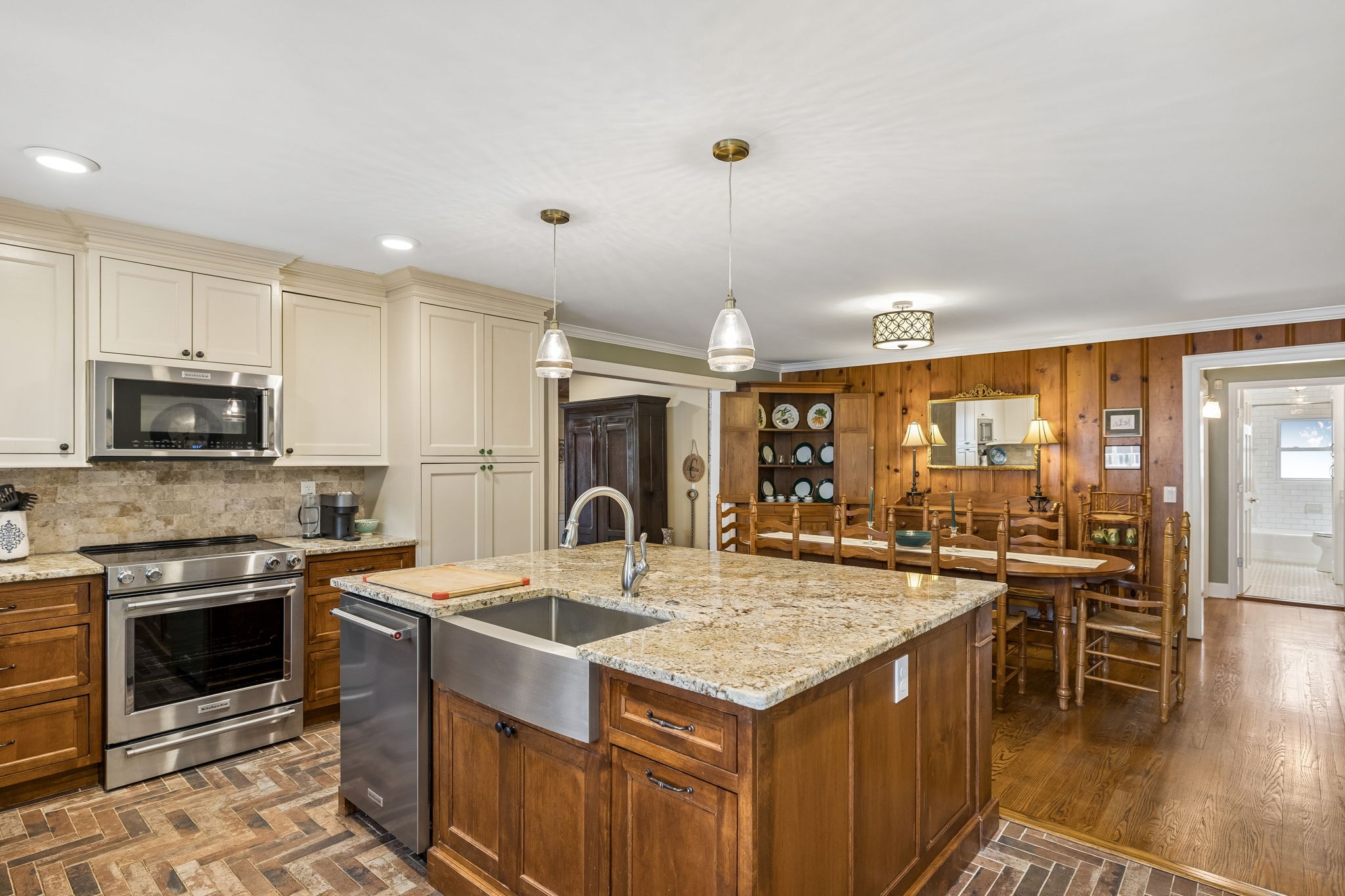 Kitchen with a large granite island, cream cabinets, stainless appliances, and an adjoining wood-paneled dining area.
