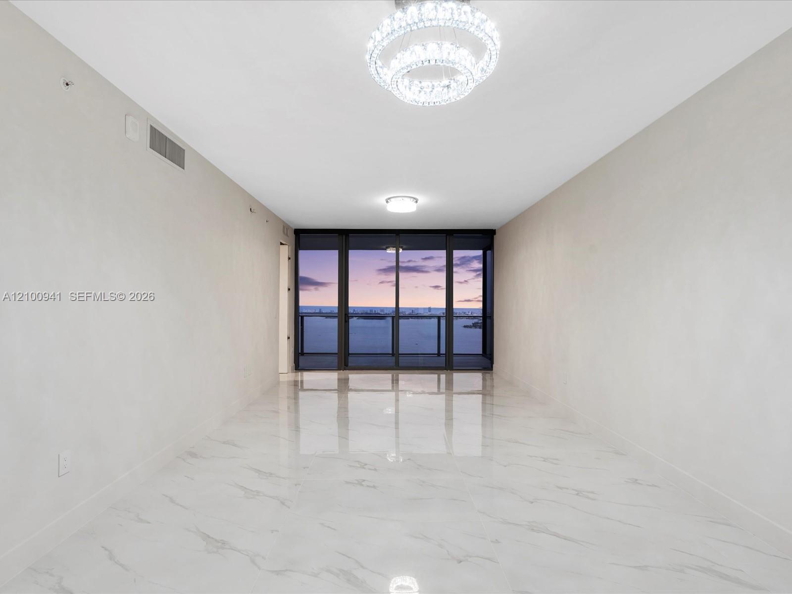 Empty condo living area with marble floors, a crystal chandelier, and dusk views over the water.