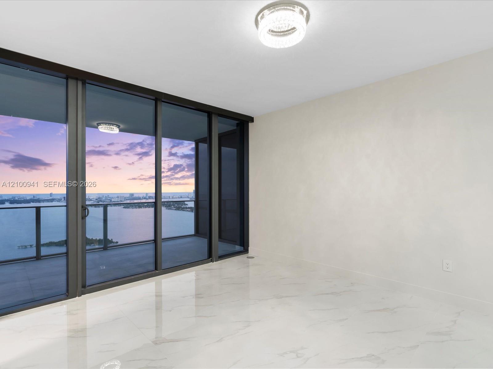 Vacant condo room with marble-look floors and floor-to-ceiling doors overlooking water and the city at dusk.