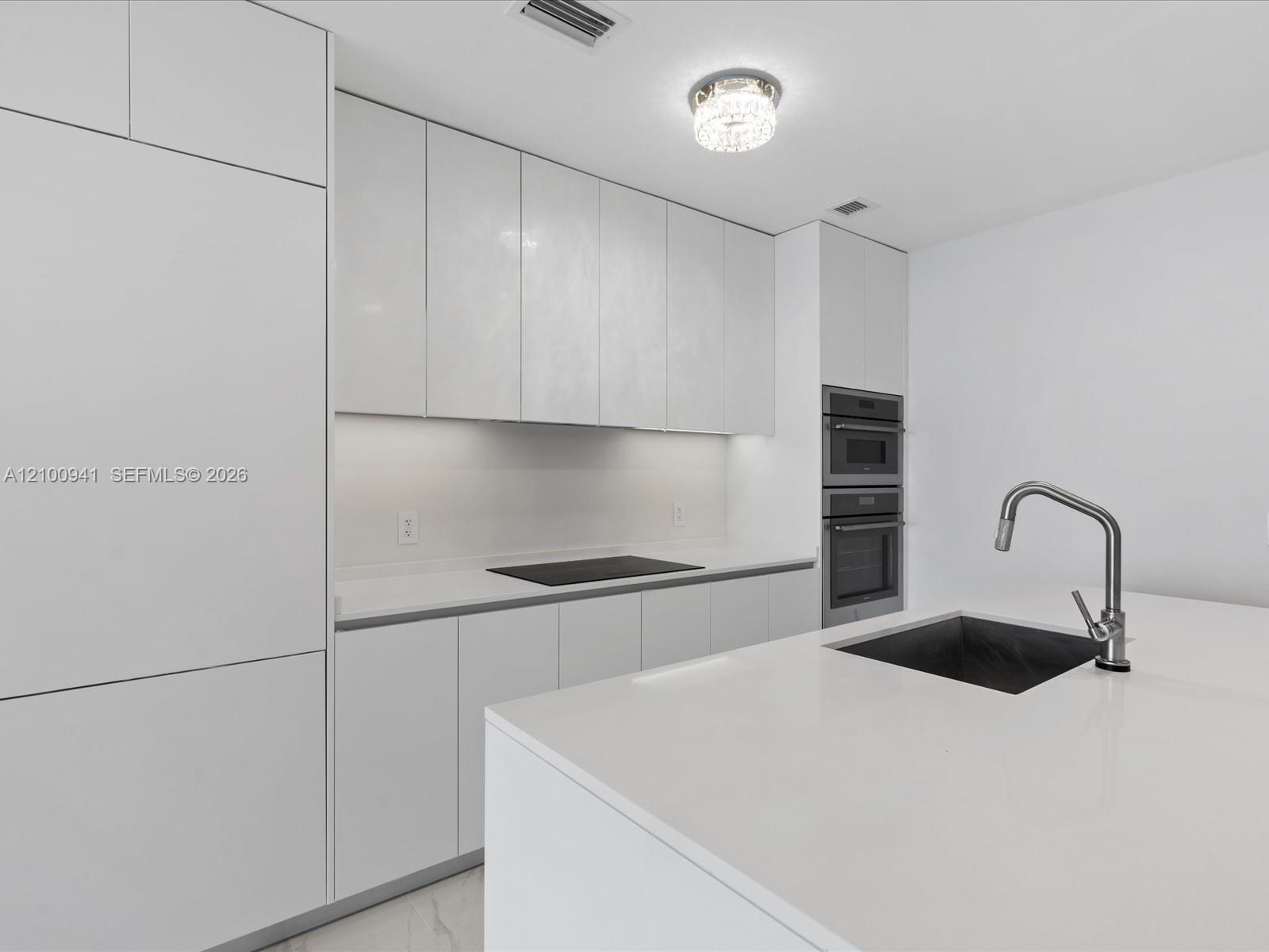 Modern white kitchen with a large island sink, induction cooktop, and stacked built-in ovens.
