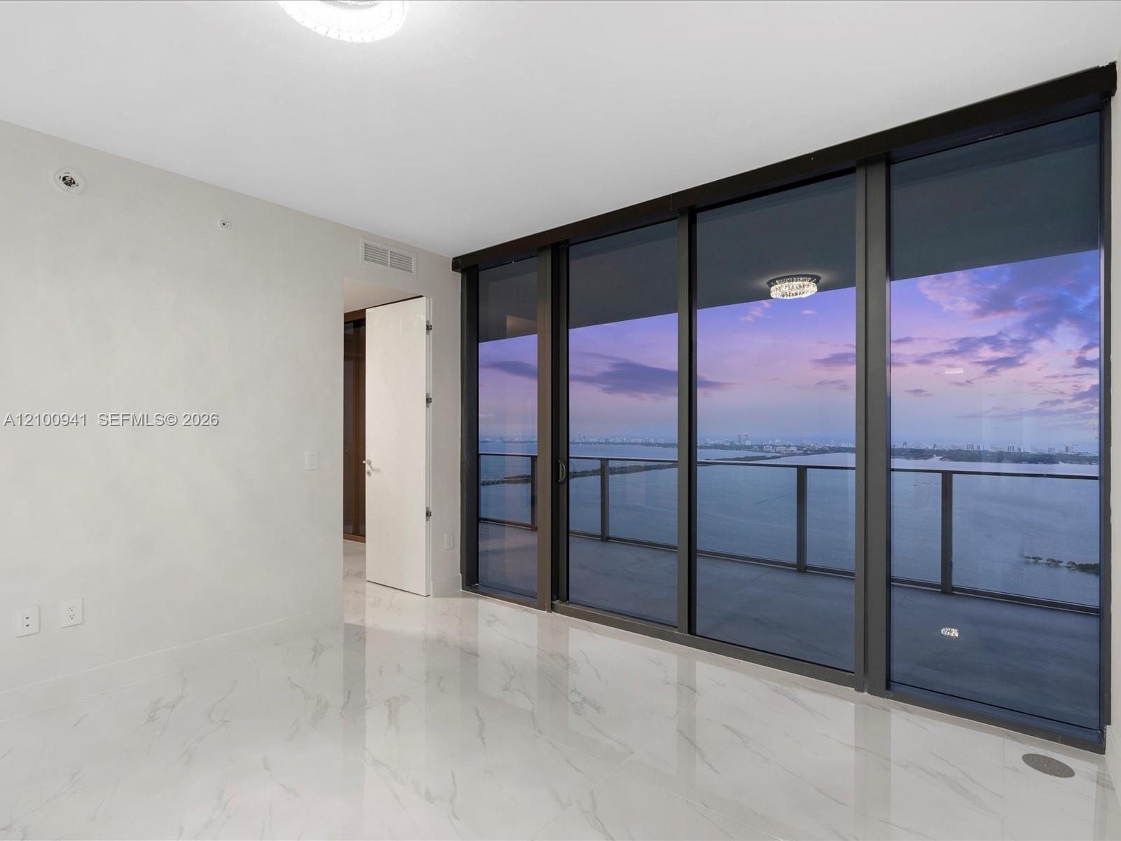 Unfurnished condo room with floor-to-ceiling glass doors overlooking water and a city skyline at dusk.