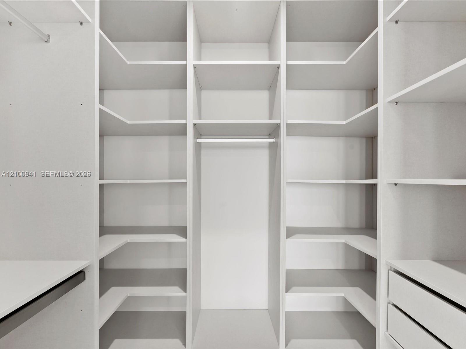 Walk-in closet with white built-in shelves, drawers, and a central hanging rod.