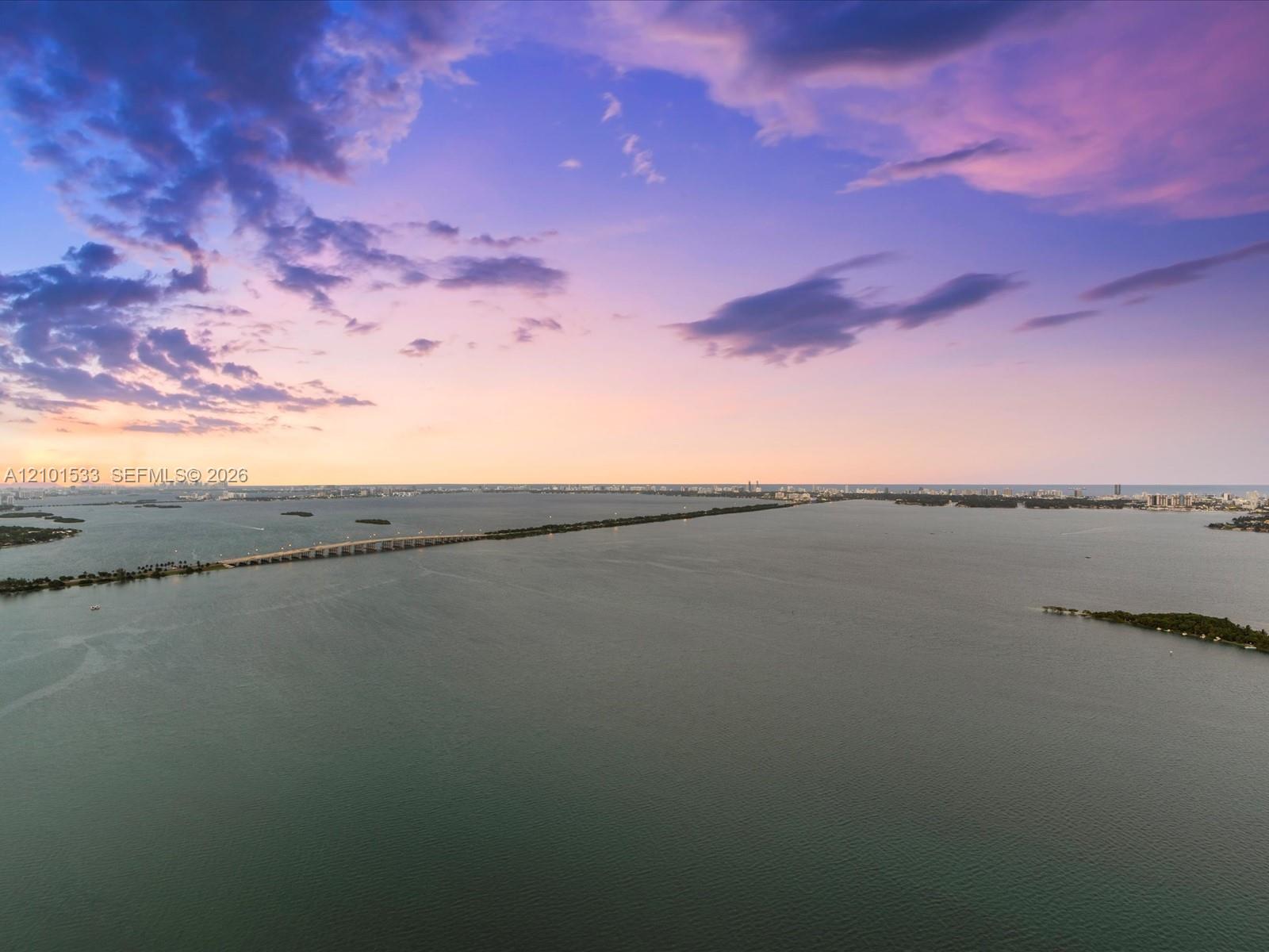 Aerial view across Biscayne Bay toward Miami at dusk, with a causeway and distant skyline.