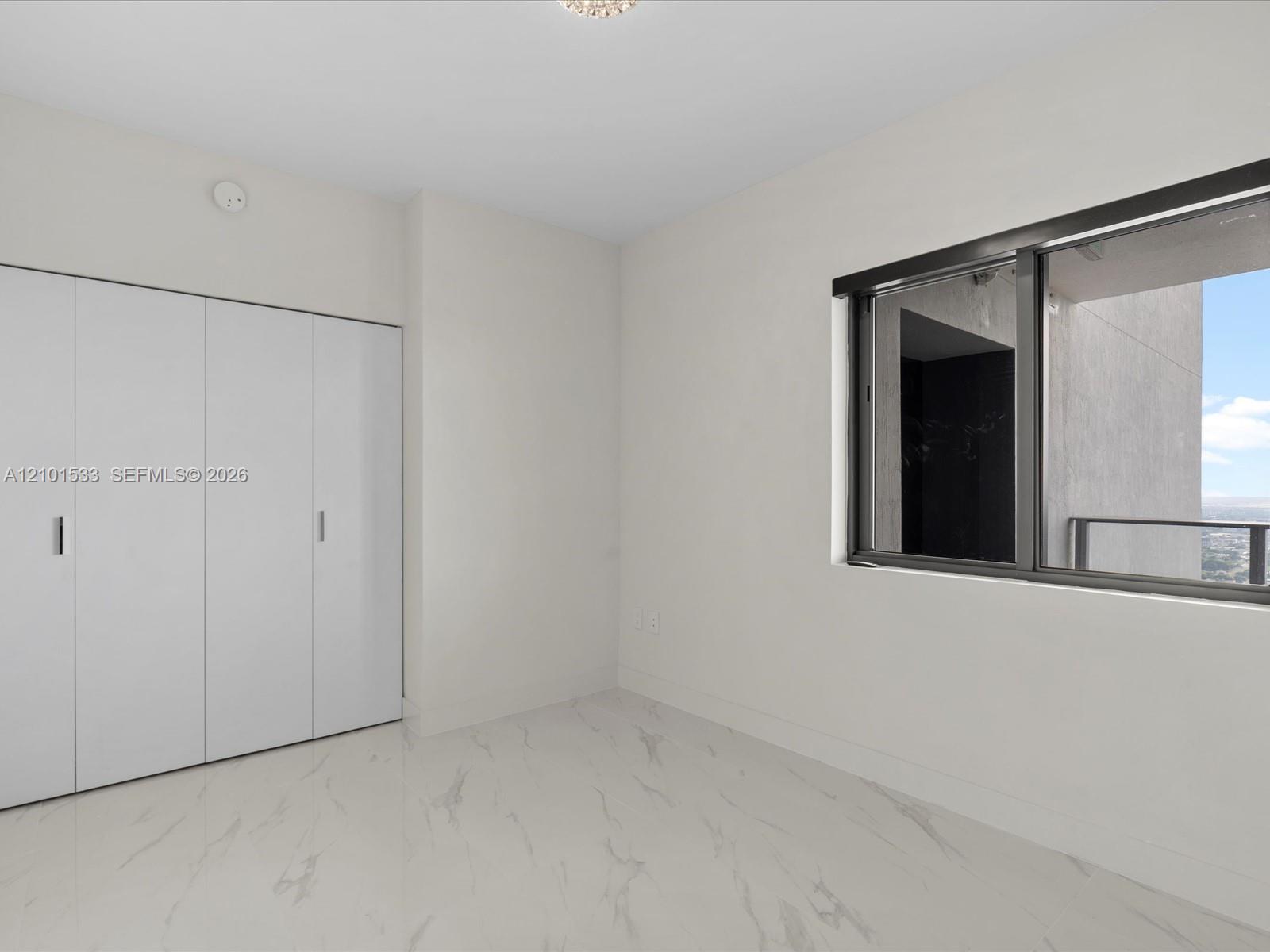 Empty condo room with built-in white closets, marble-look flooring, and a balcony window.