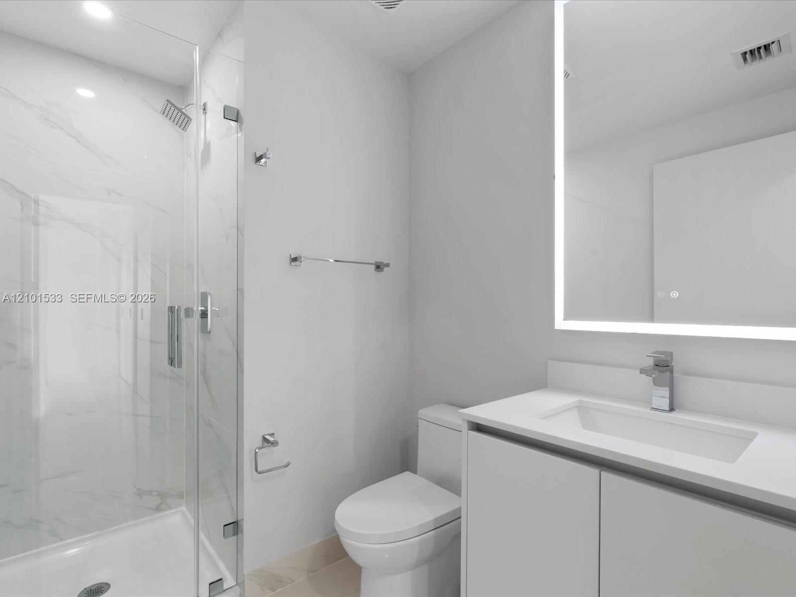 Modern bathroom with a glass shower, marble-look tile, white vanity, and illuminated mirror.