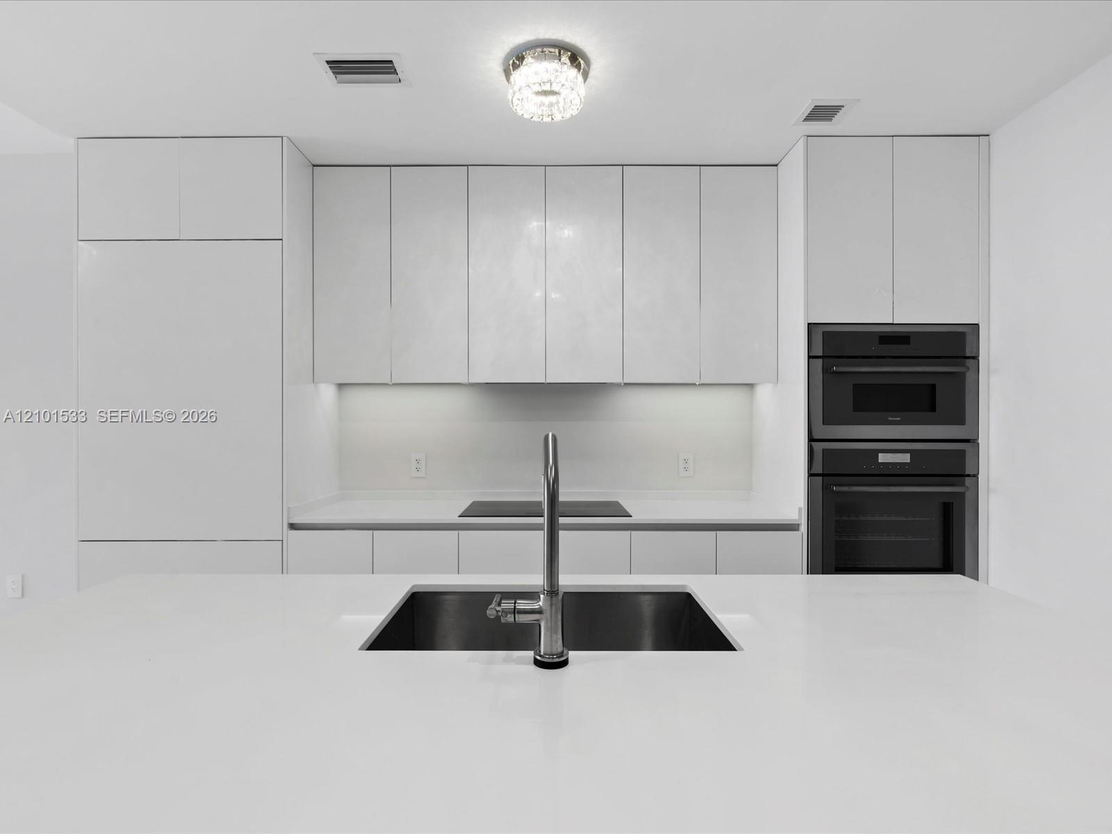 Modern white kitchen with a large island sink, induction cooktop, and stacked built-in ovens.