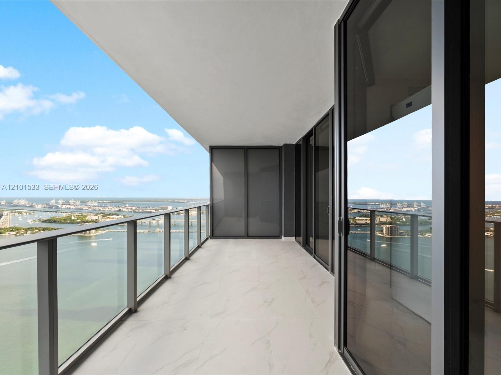 High-rise balcony with glass railing and marble-look tile overlooking water and distant port cranes.