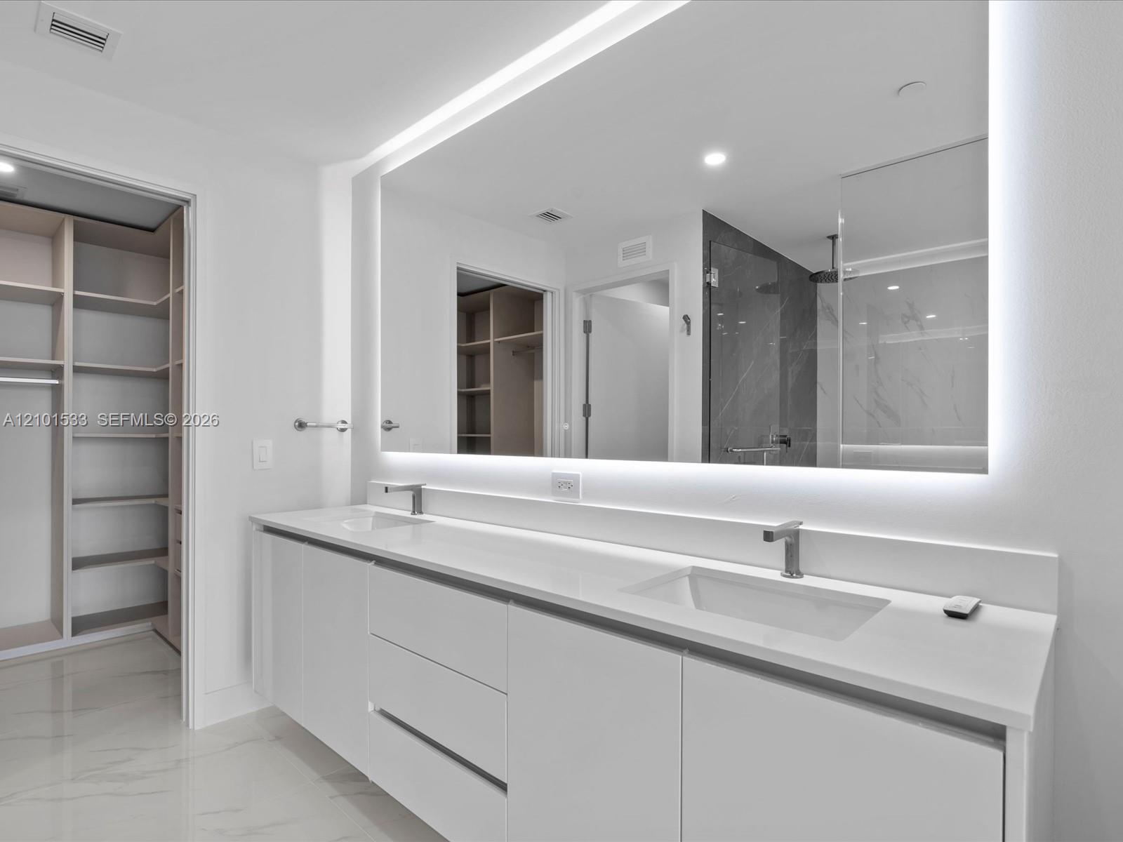Modern bathroom with a double-sink vanity, illuminated mirror, glass shower, and open walk-in closet.