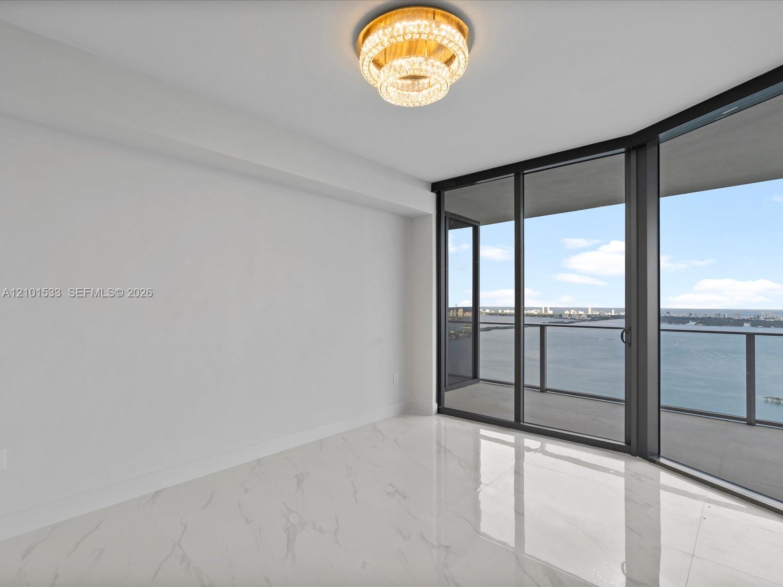 Vacant condo room with marble-look floors, a chandelier, and balcony views over the water and city.