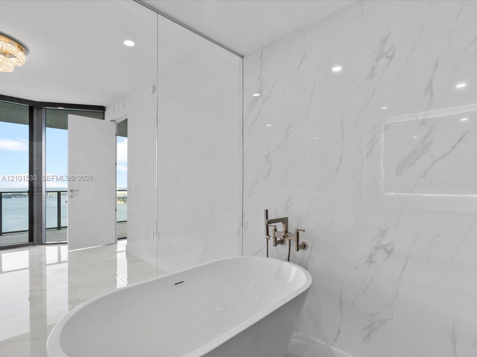 Modern bathroom with a freestanding tub beside marble-look tile and an open doorway overlooking the water.