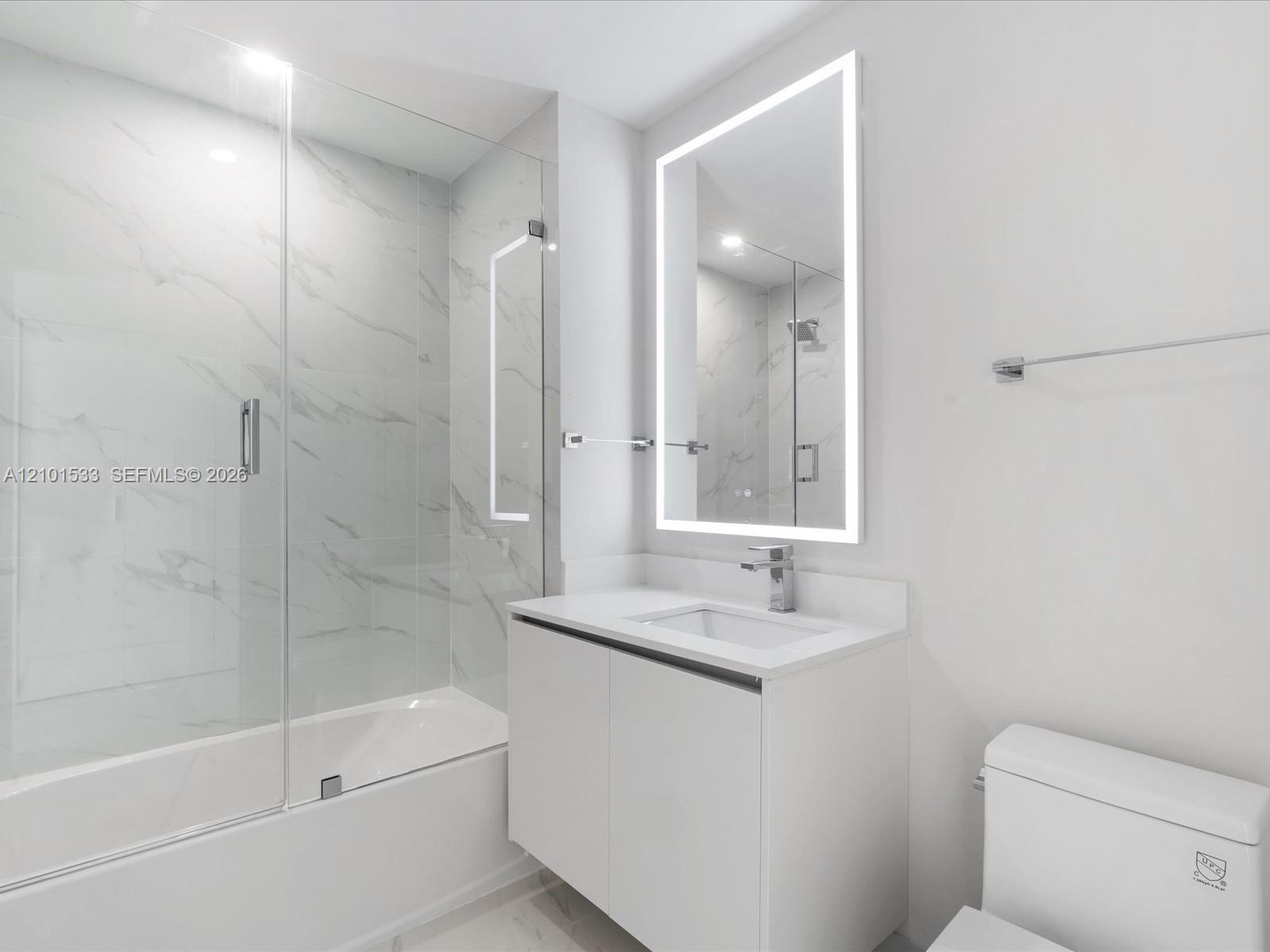 Modern bathroom with a glass-enclosed tub, marble-look tile, floating vanity, and illuminated mirror.