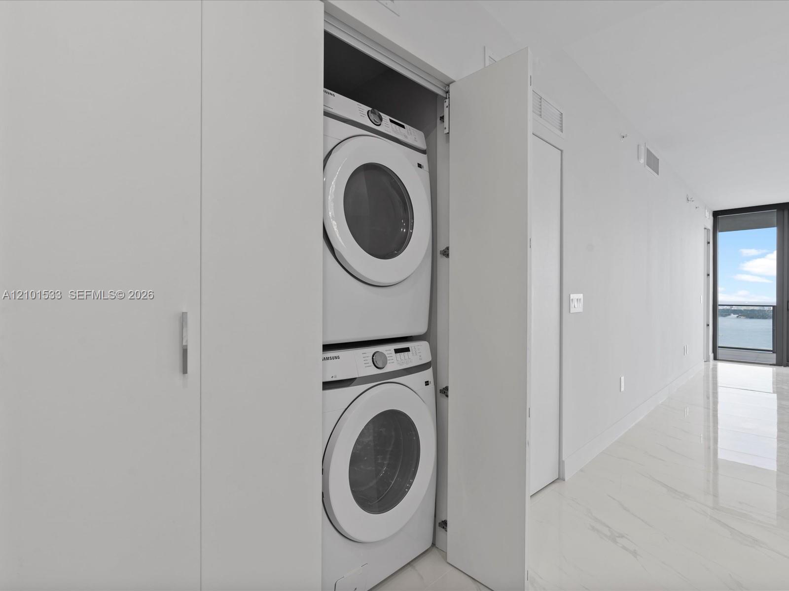 Stacked washer and dryer in a hallway laundry closet, with a water view through the balcony window.