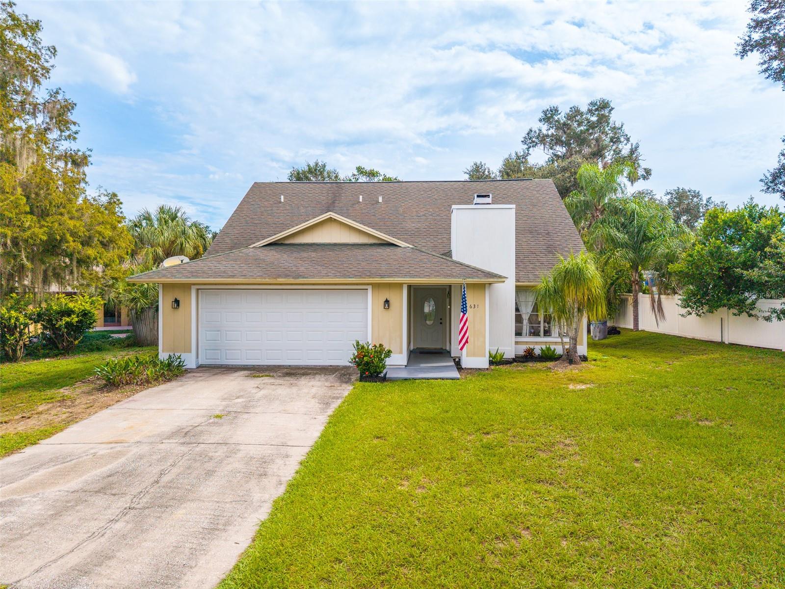 Two-story beige home with a double garage, American flag by the entry, and a broad front lawn.
