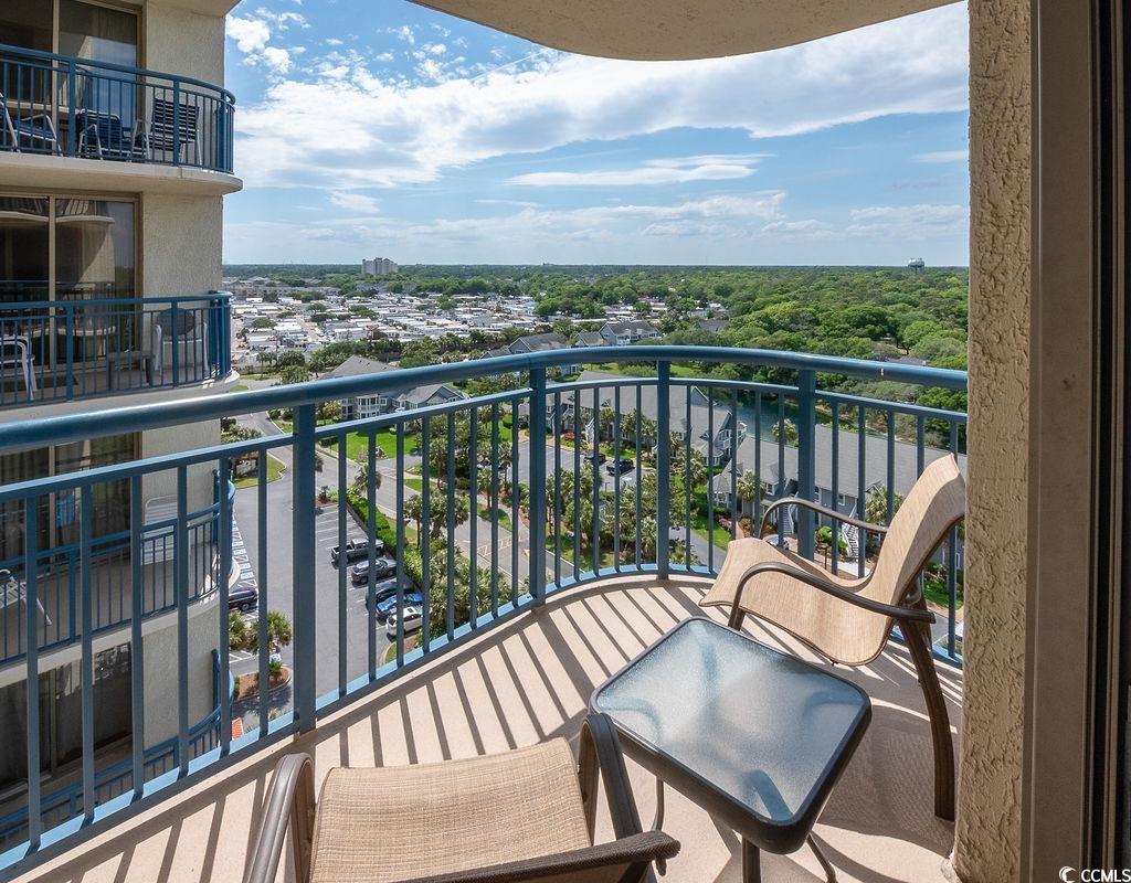 High-rise balcony with blue railings and outdoor seating overlooking rooftops and trees.