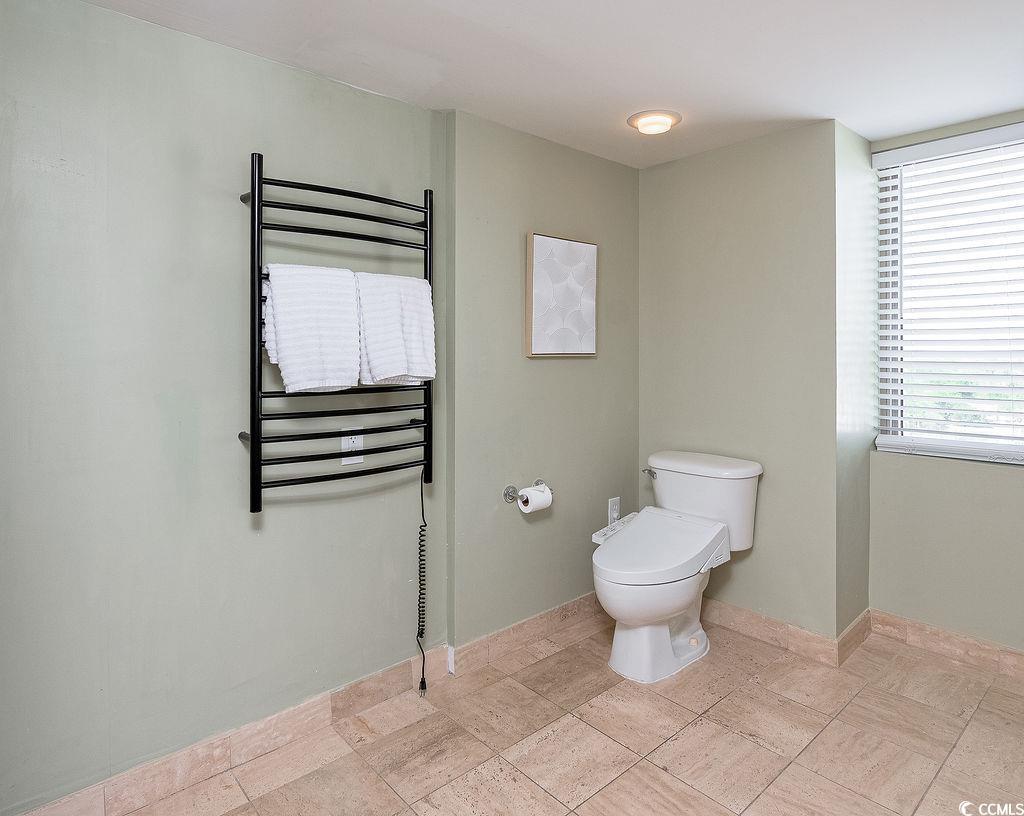 Bathroom with a heated towel rack holding white towels beside a toilet and window with blinds.