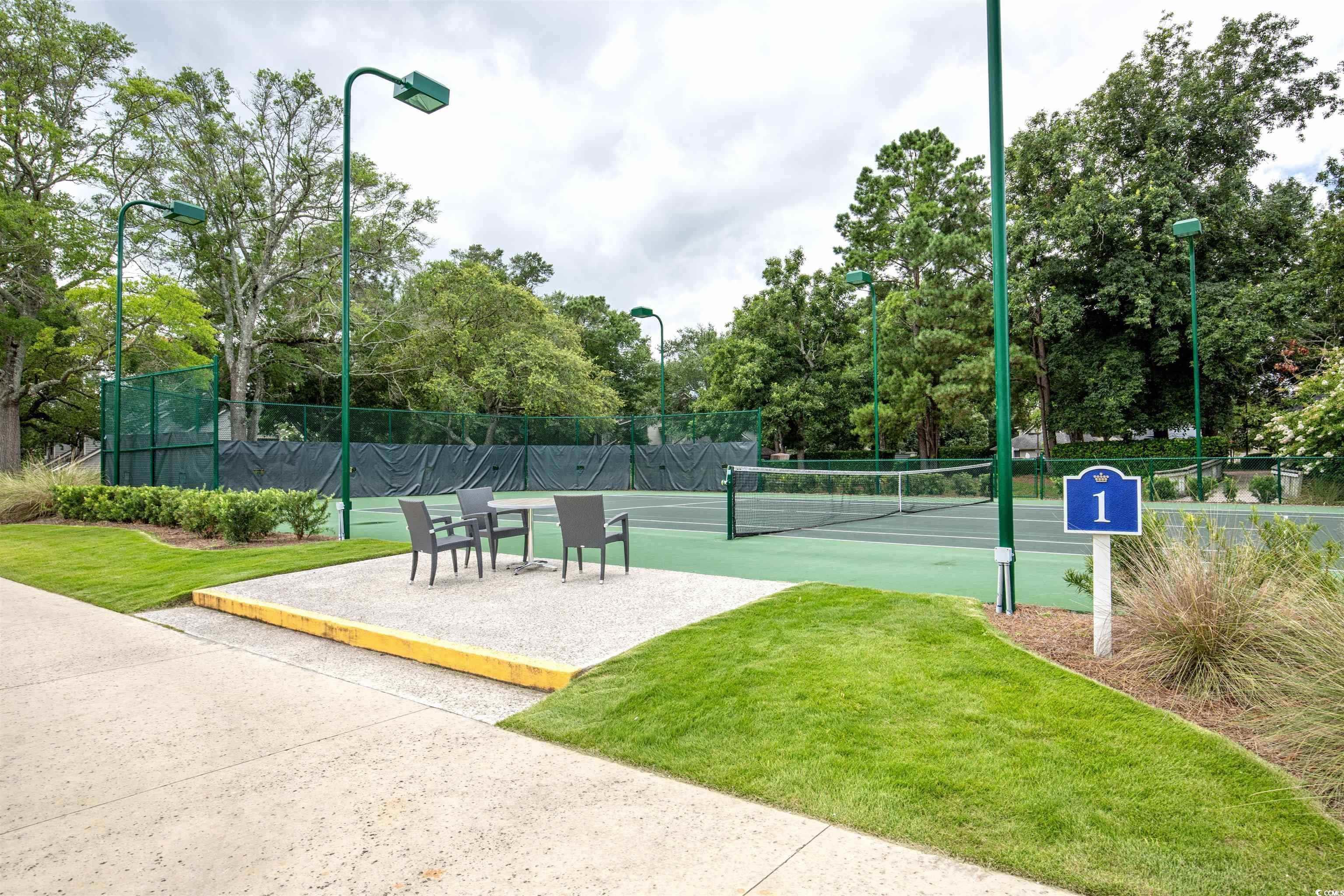 Outdoor tennis court with green fencing, tall lights, and a patio seating area.