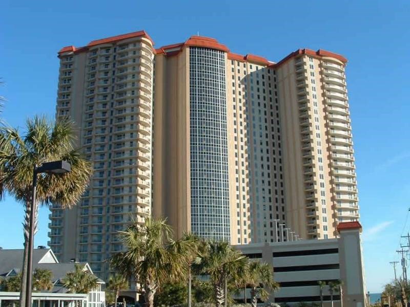 Beige high-rise condominium with curved balconies, a central glass facade, and palm trees in front.