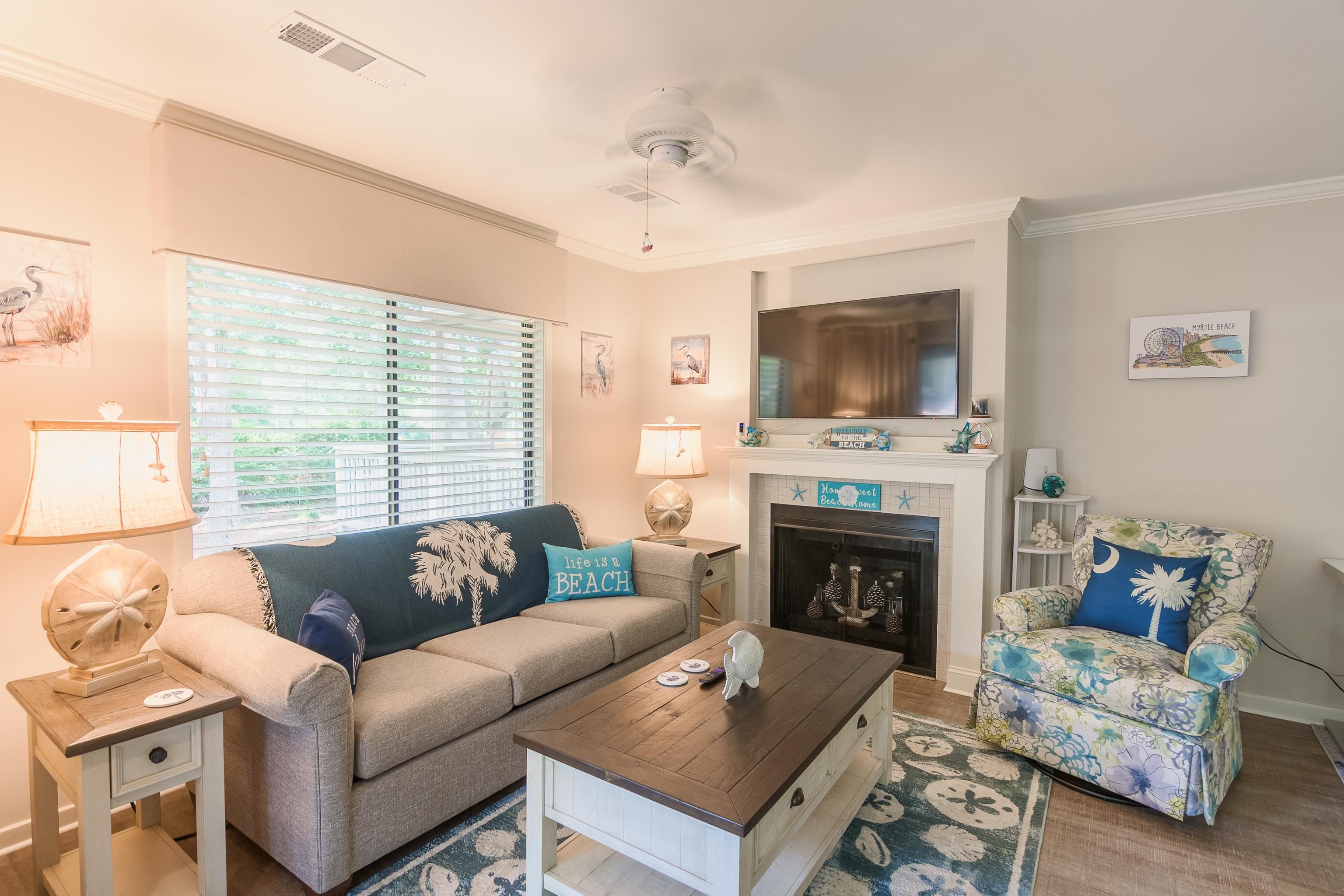 Furnished living room with beach-themed décor, a fireplace, and large windows with blinds.