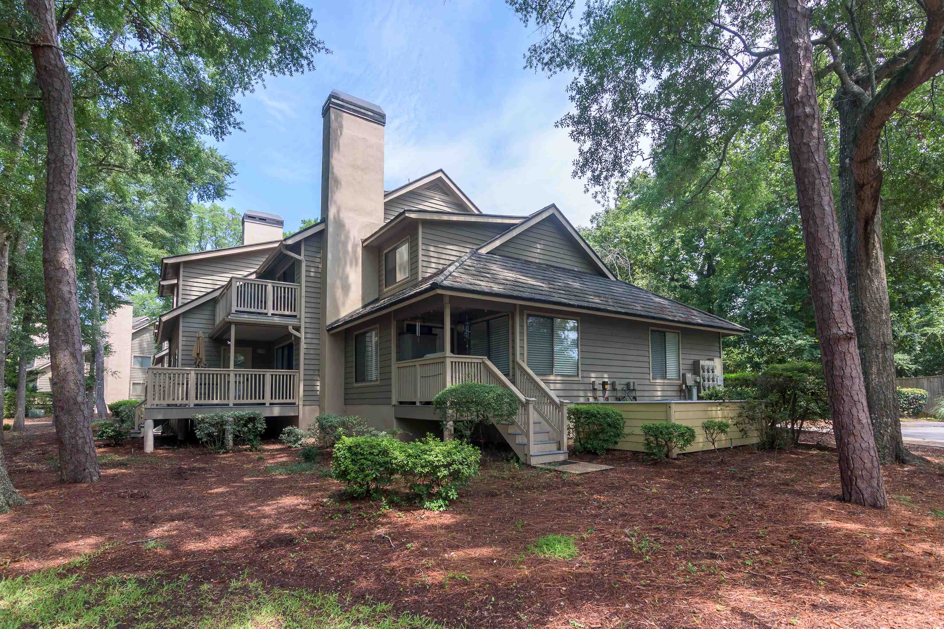 Gray-sided home with raised decks and tall chimneys among mature trees.