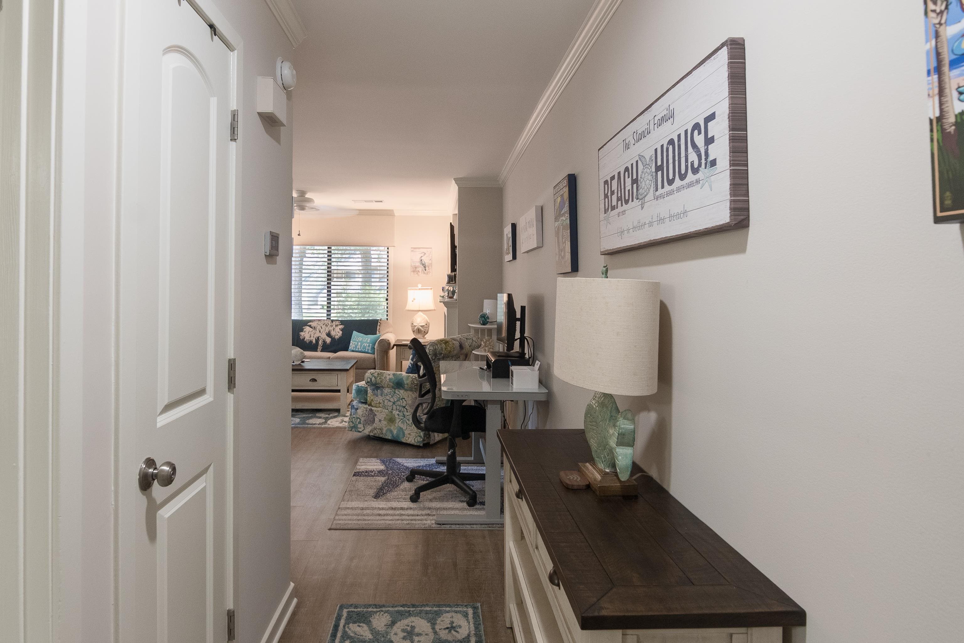 Furnished condo hallway opens to a living room, with a desk and beach-themed wall sign.
