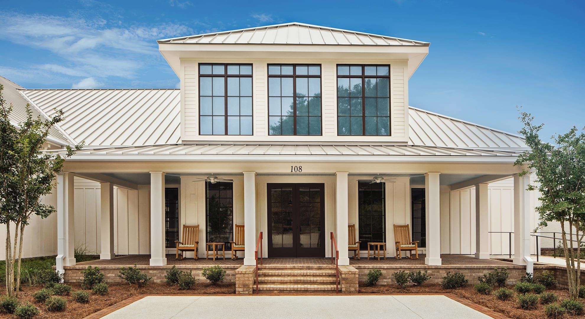 White building entrance with a broad covered porch, metal roof, and wooden chairs.