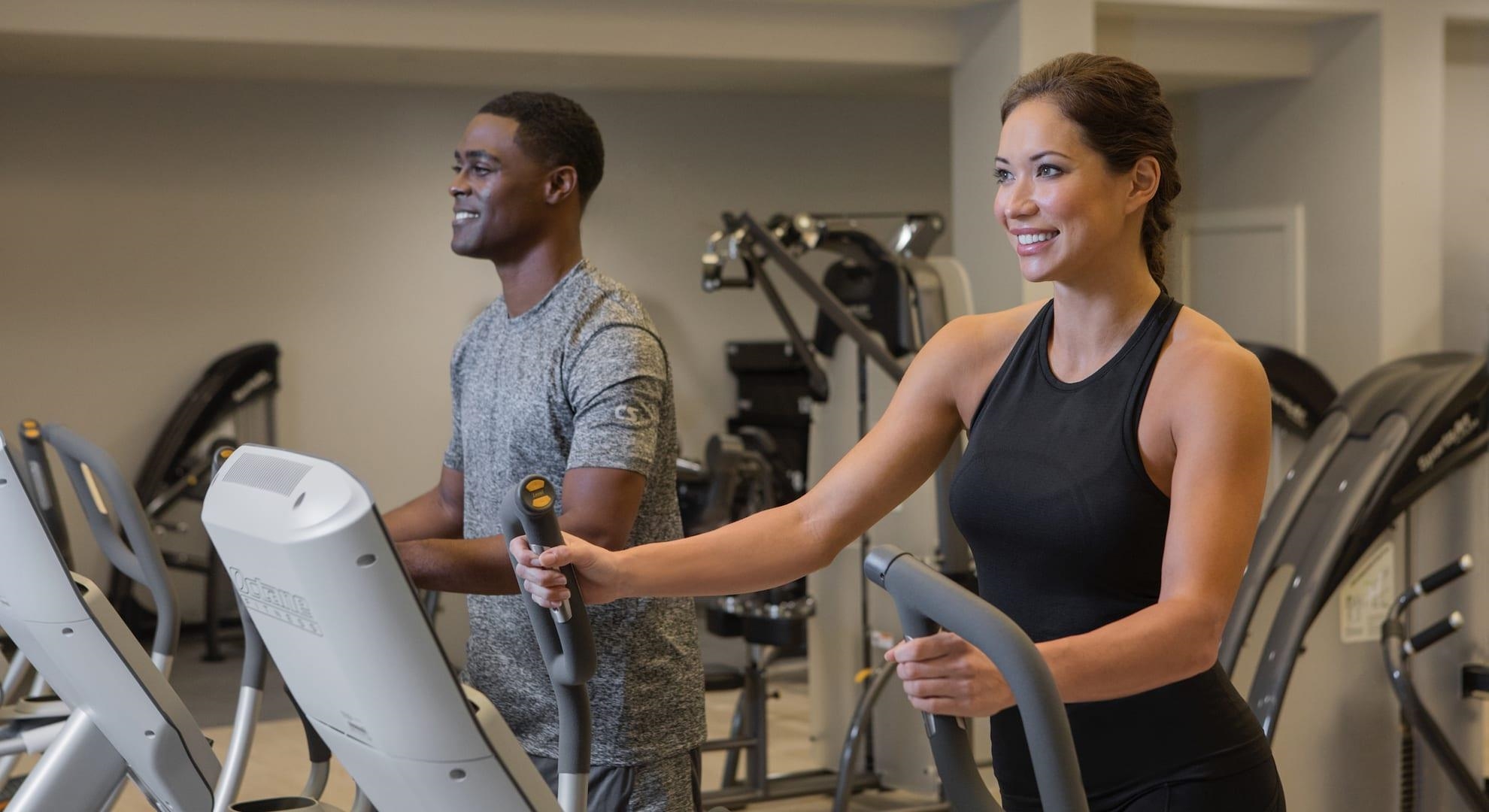 Two people exercise on elliptical machines in a gym.
