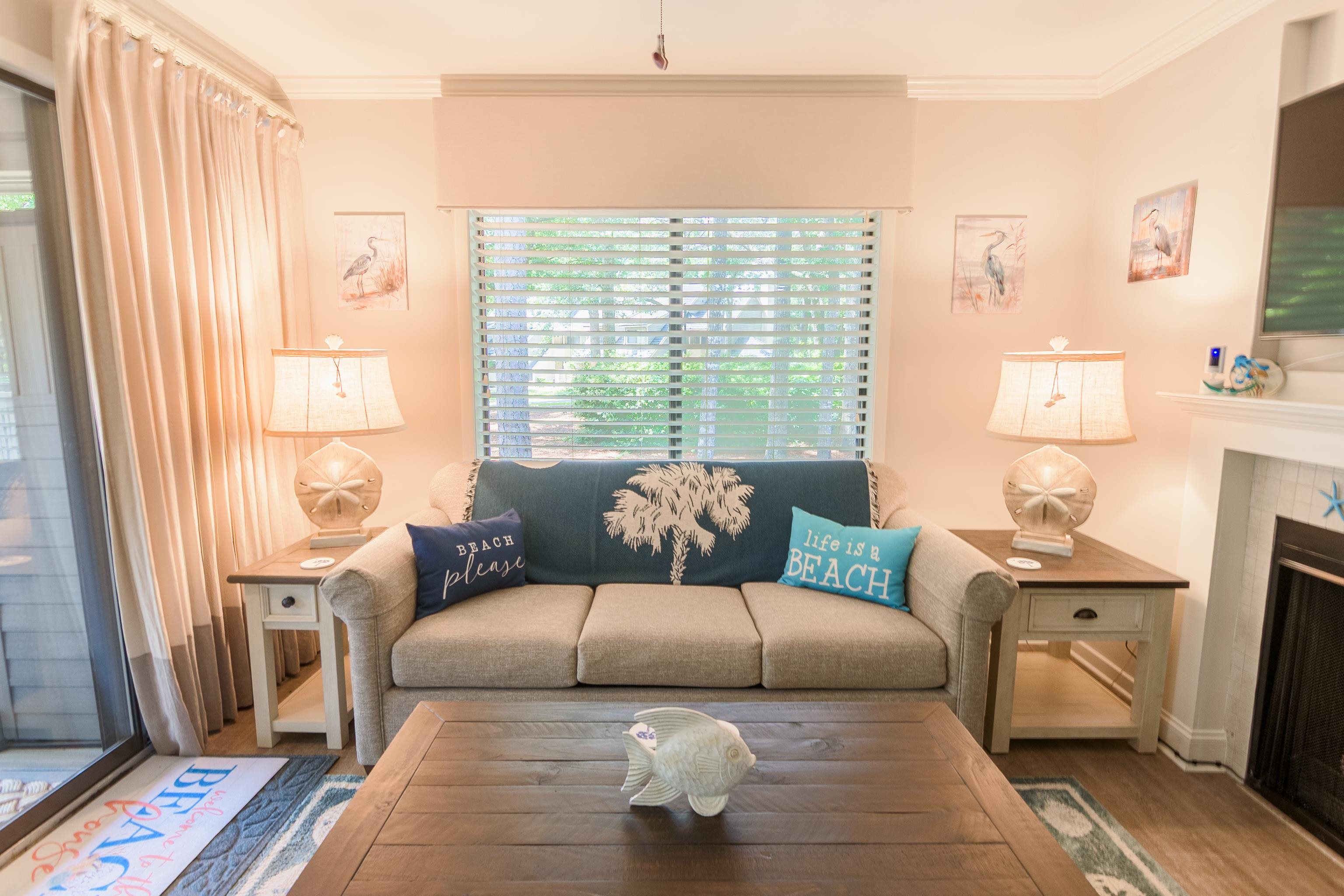 Furnished living room with a gray sofa, beach-themed pillows, and a coffee table beside a fireplace.