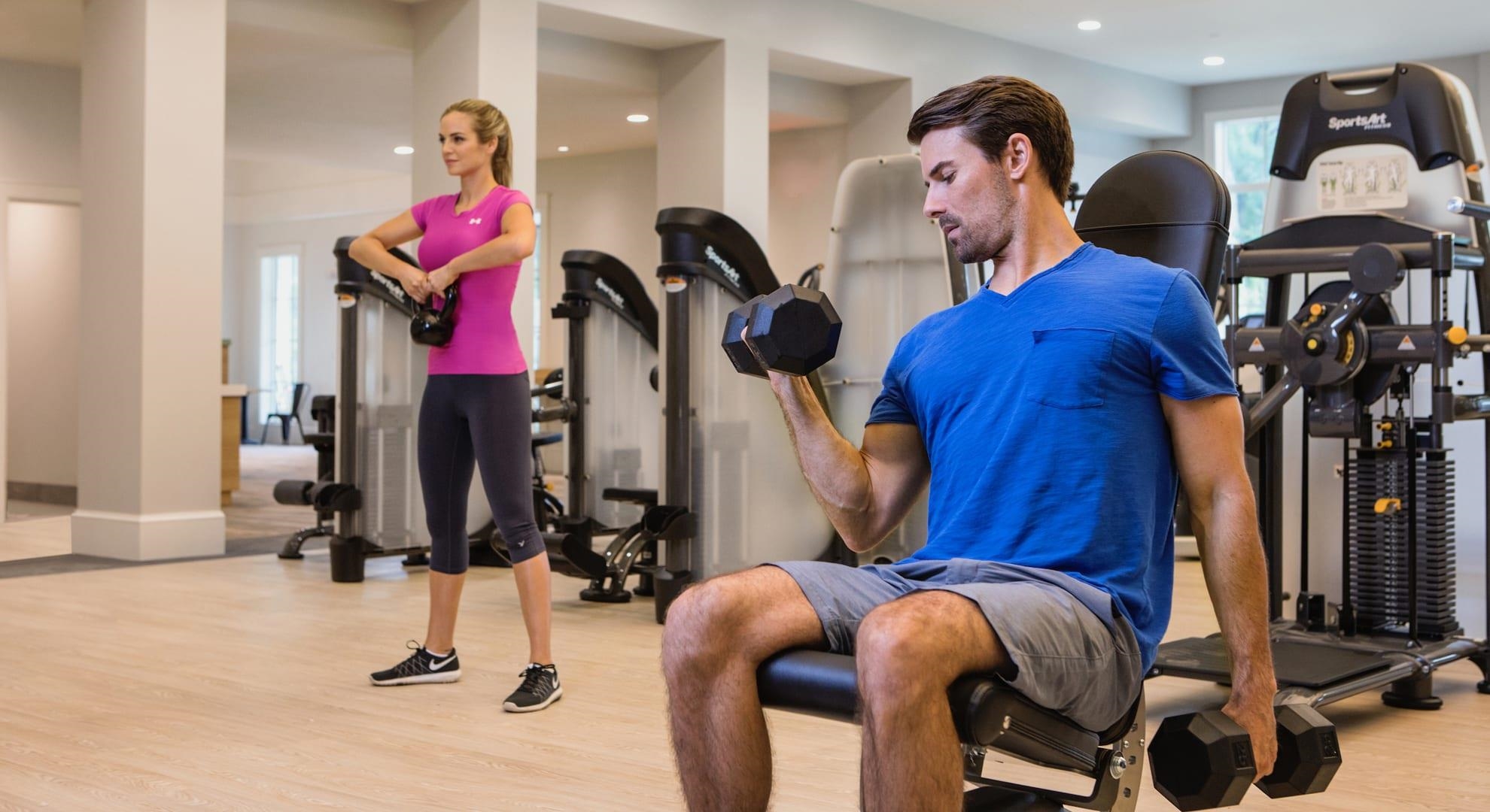 Fitness center with weight machines and two people exercising, including a seated man lifting a dumbbell.