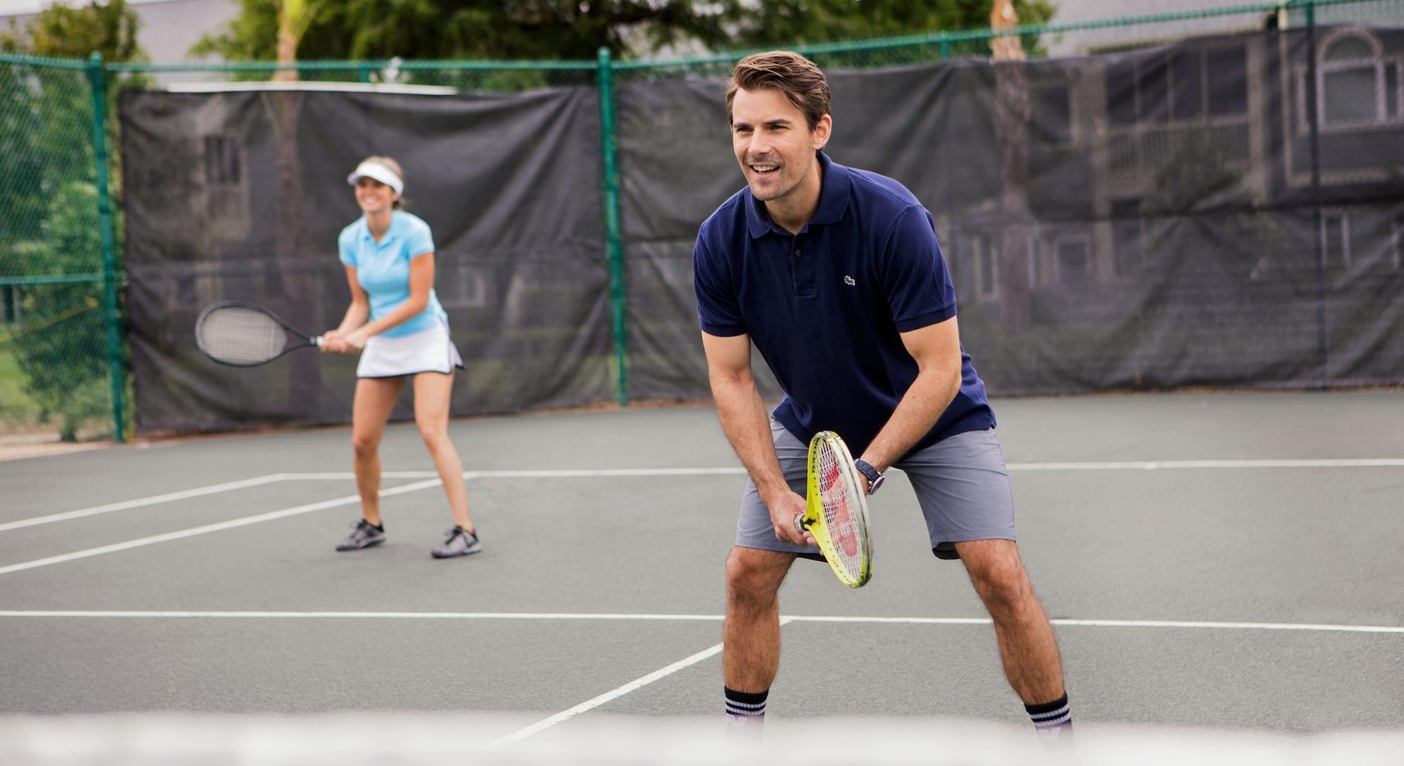 Two adults play tennis on an outdoor court, with the man in the foreground holding a racket.