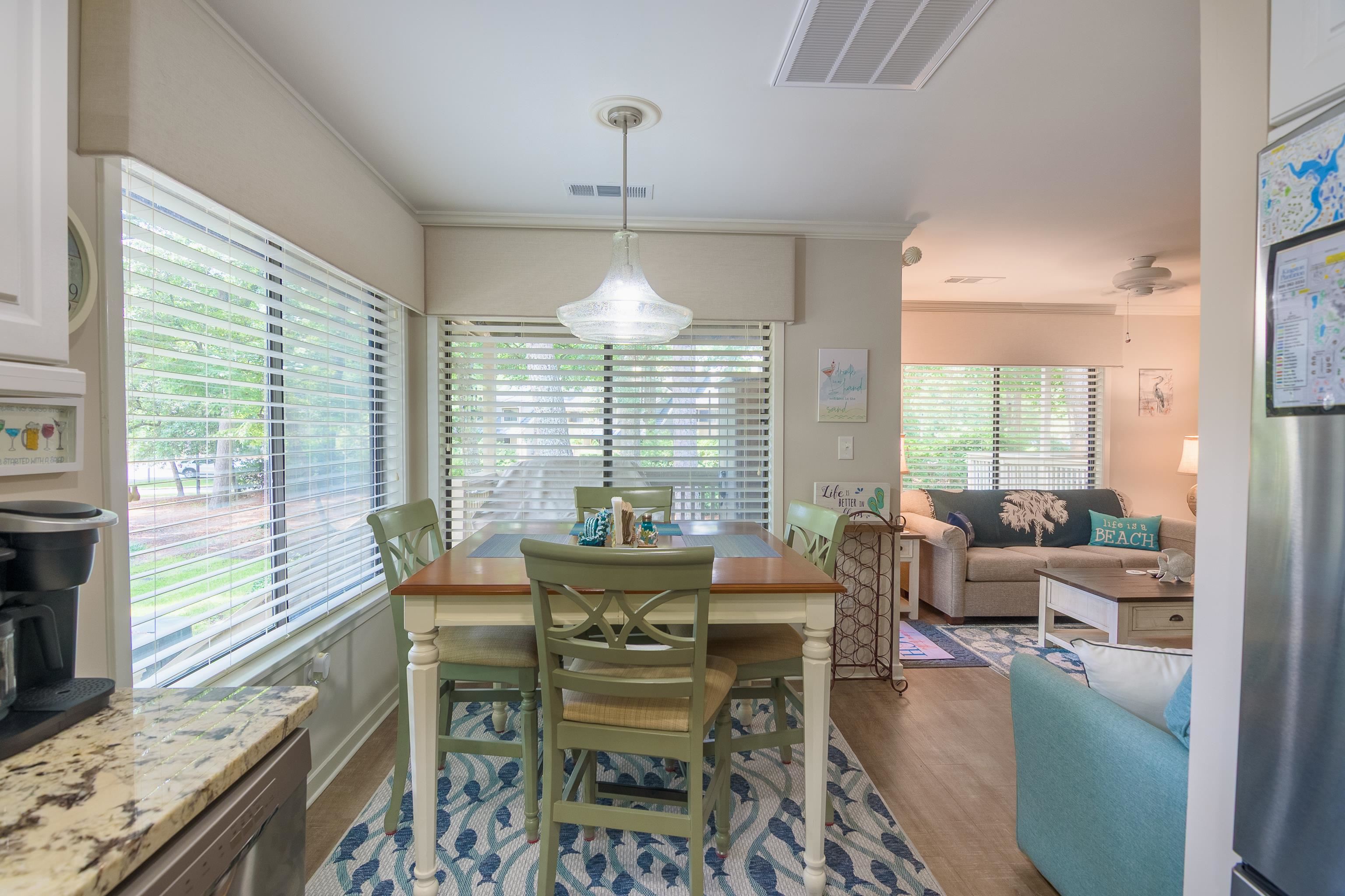 Dining area with green chairs and a patterned rug, opening onto a beach-themed living room.