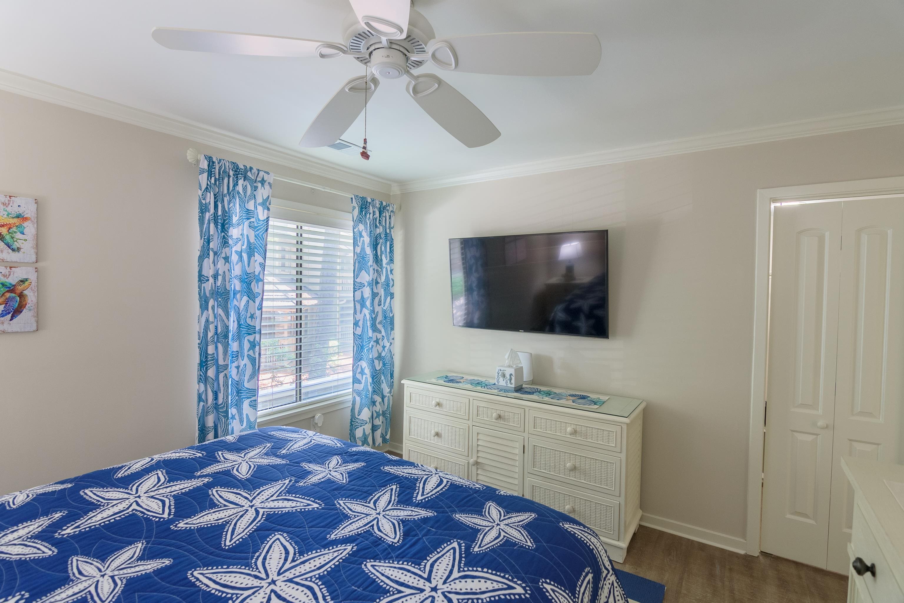 Bedroom with blue starfish-patterned bedding, a wall-mounted TV, and a window with blue curtains.