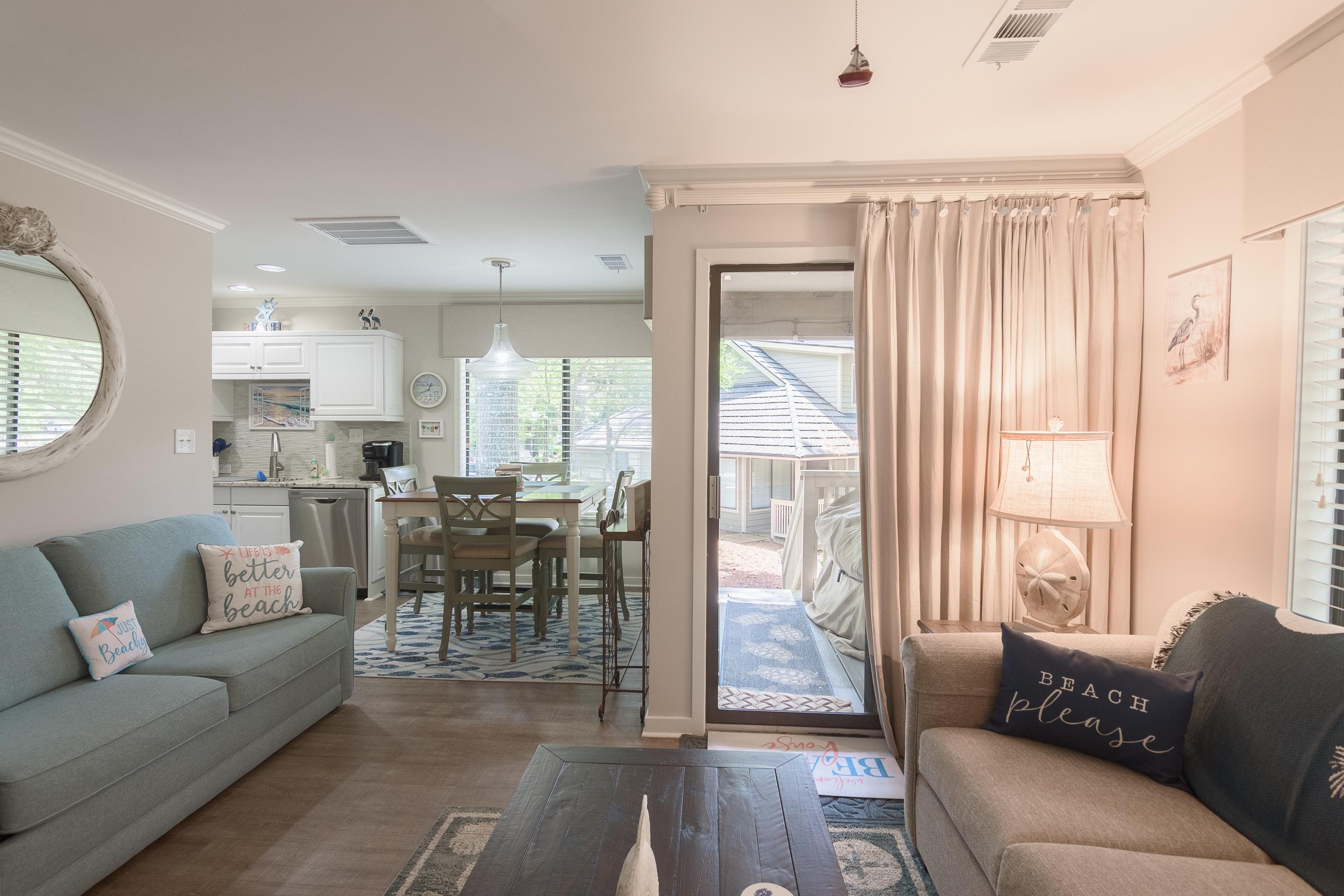 Furnished living room opening to a dining area and kitchen, with a sliding glass door to the patio.