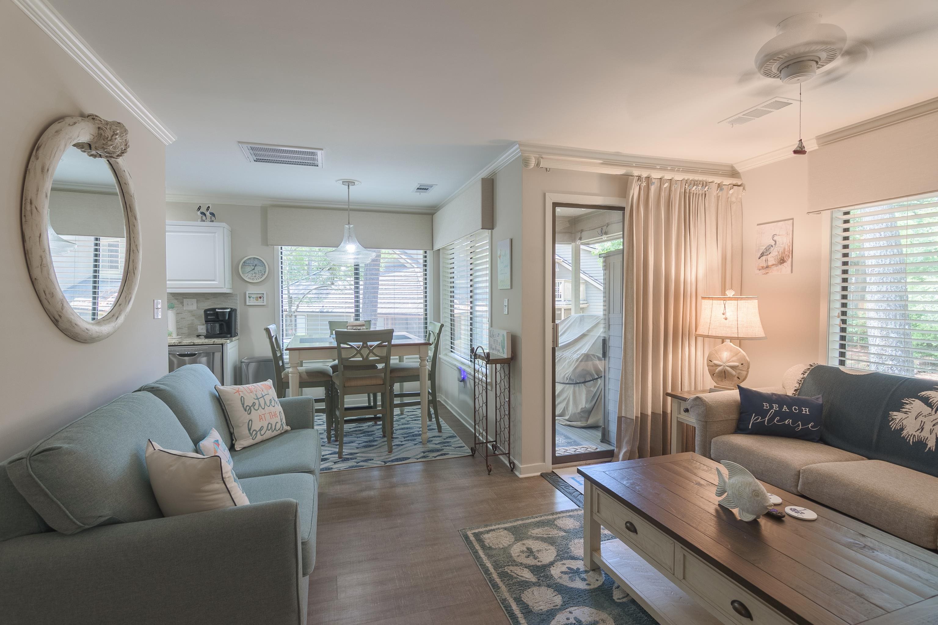 Furnished living room opening to a dining area and compact kitchen, with beach-themed decor.