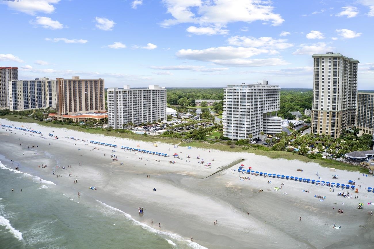 Aerial view of beachfront condominium towers along a wide sandy beach with rows of blue umbrellas.