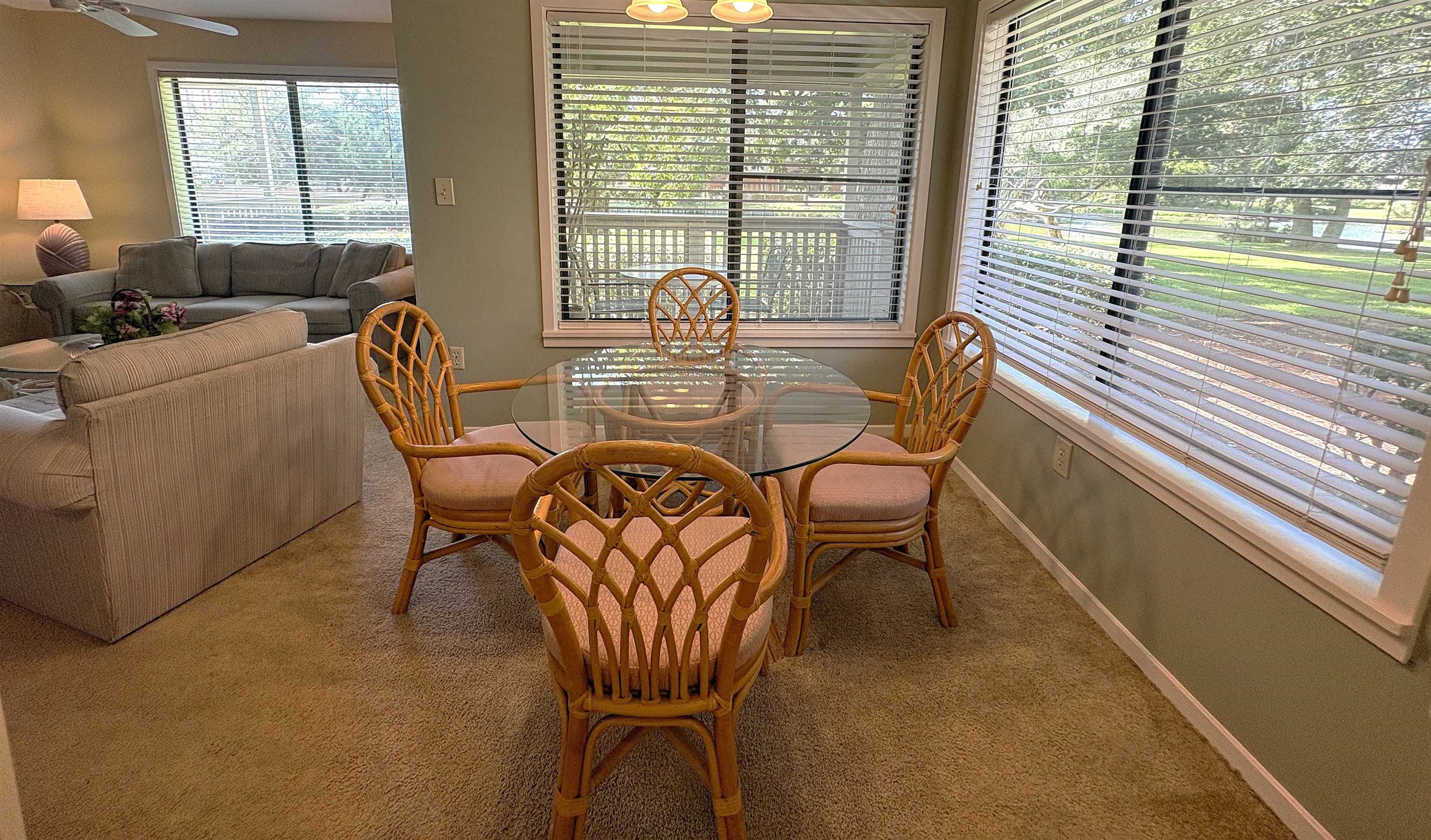 Dining area with a glass-top table and rattan chairs beside large windows overlooking greenery.
