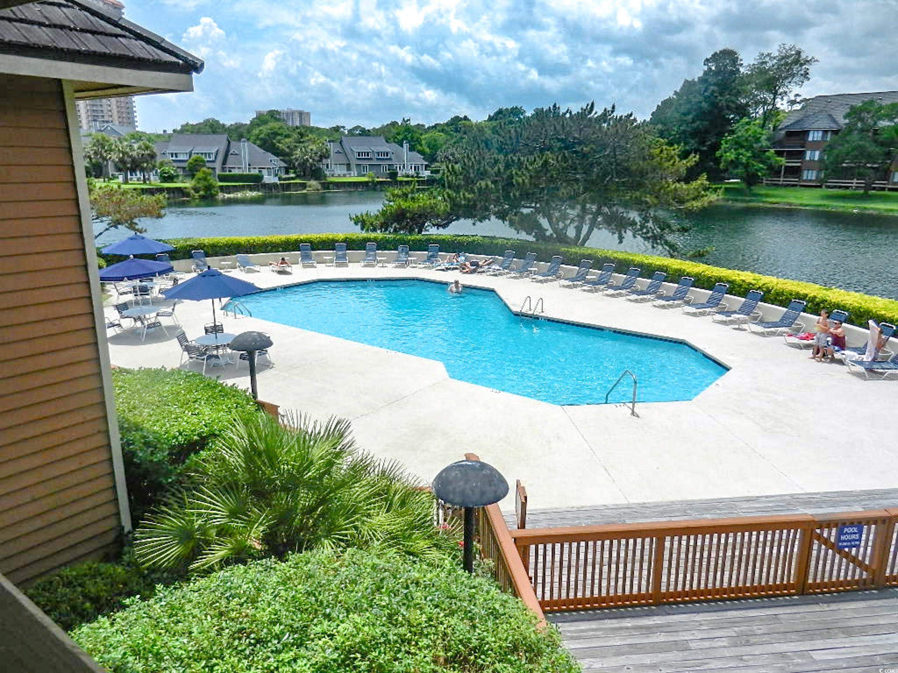 Community swimming pool beside a lake, with lounge chairs, umbrellas, and surrounding homes.