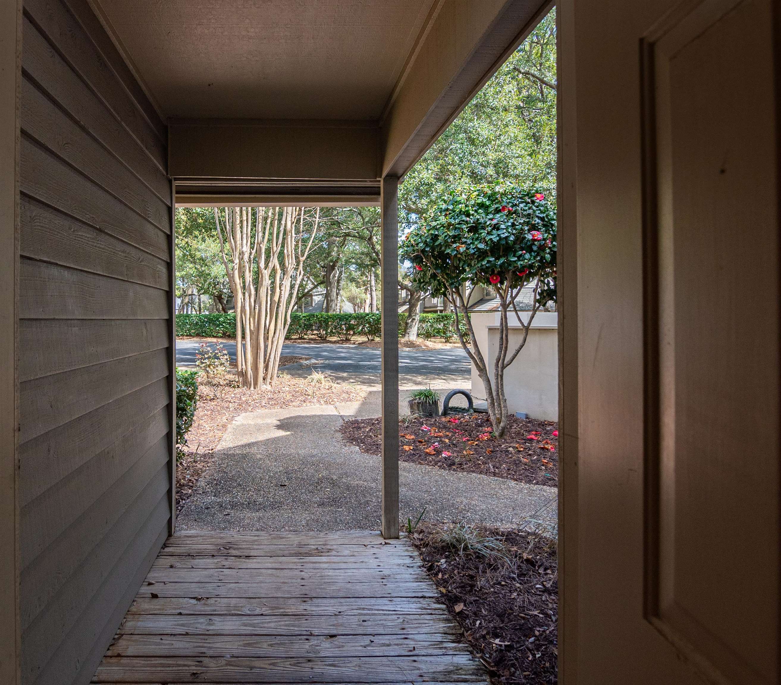Covered wooden entry porch looking out toward flowering shrubs, trees, and a walkway.