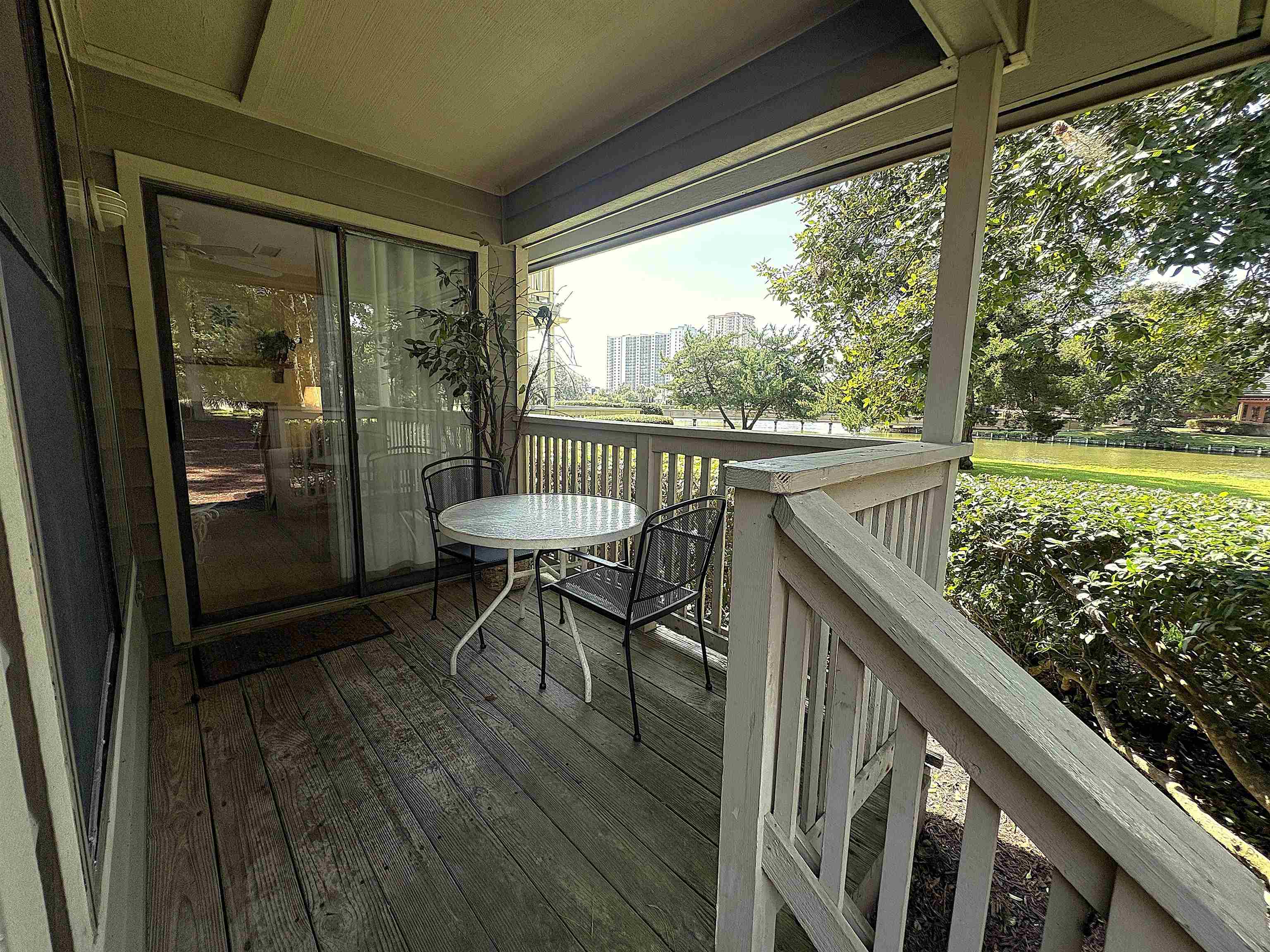 Covered condo balcony with a small table and chairs overlooking a pond and landscaped grounds.