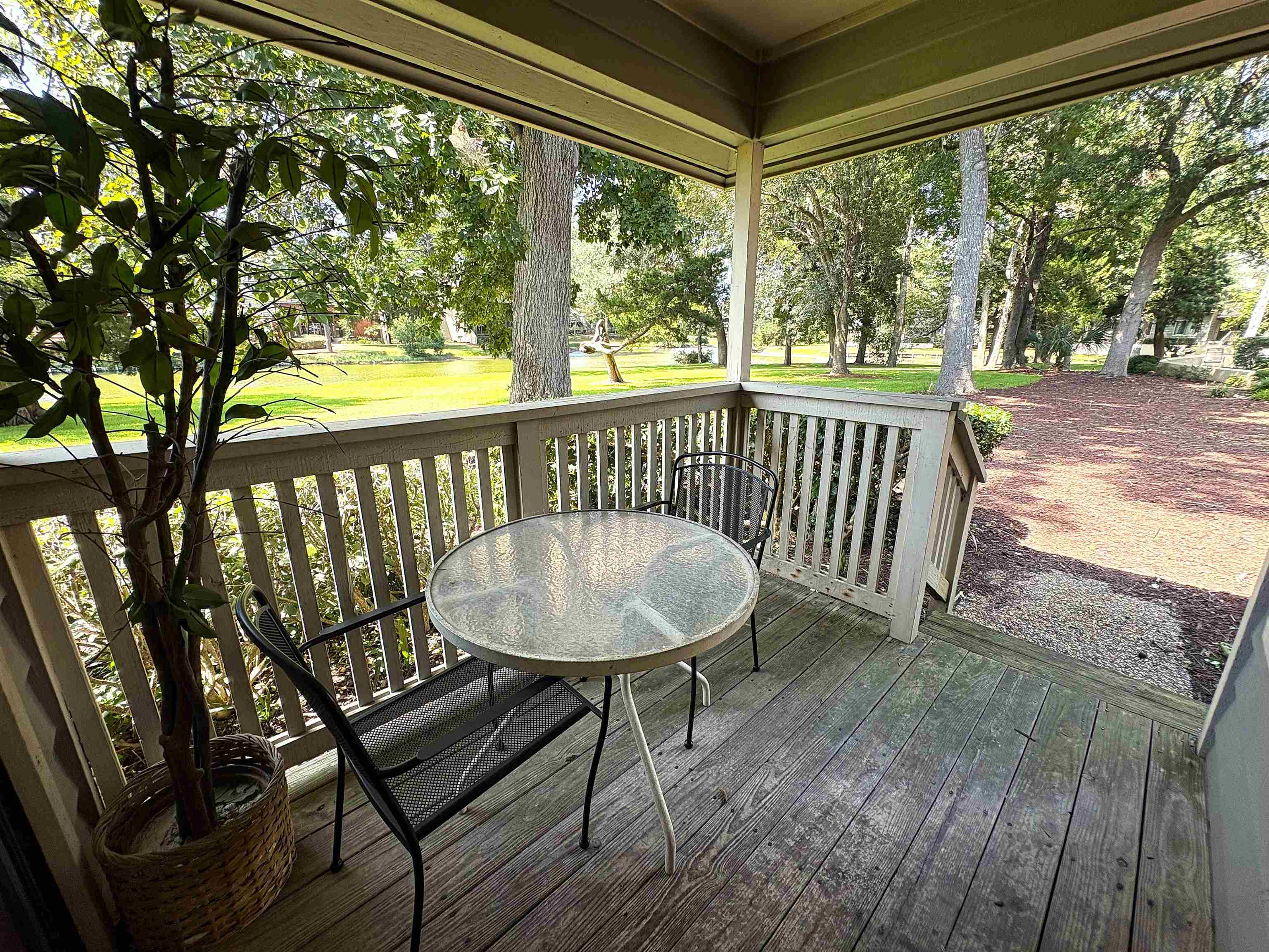 Covered wooden porch with a round glass table and chairs overlooking a leafy lawn.