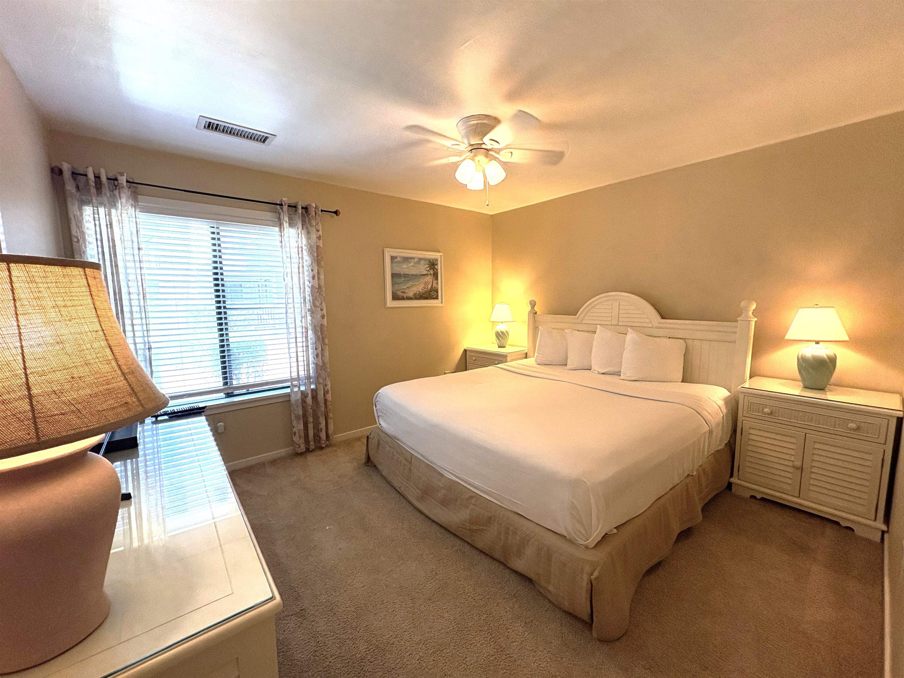 Furnished bedroom with a white king bed, matching nightstands, and a large window with blinds.
