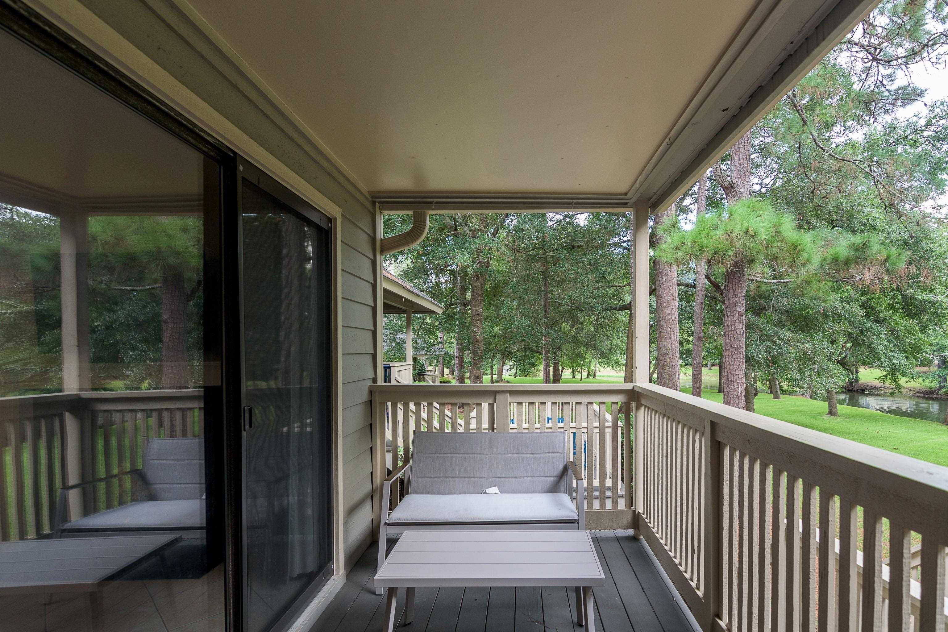 Covered balcony with gray lounge furniture overlooking a tree-lined lawn and pond.