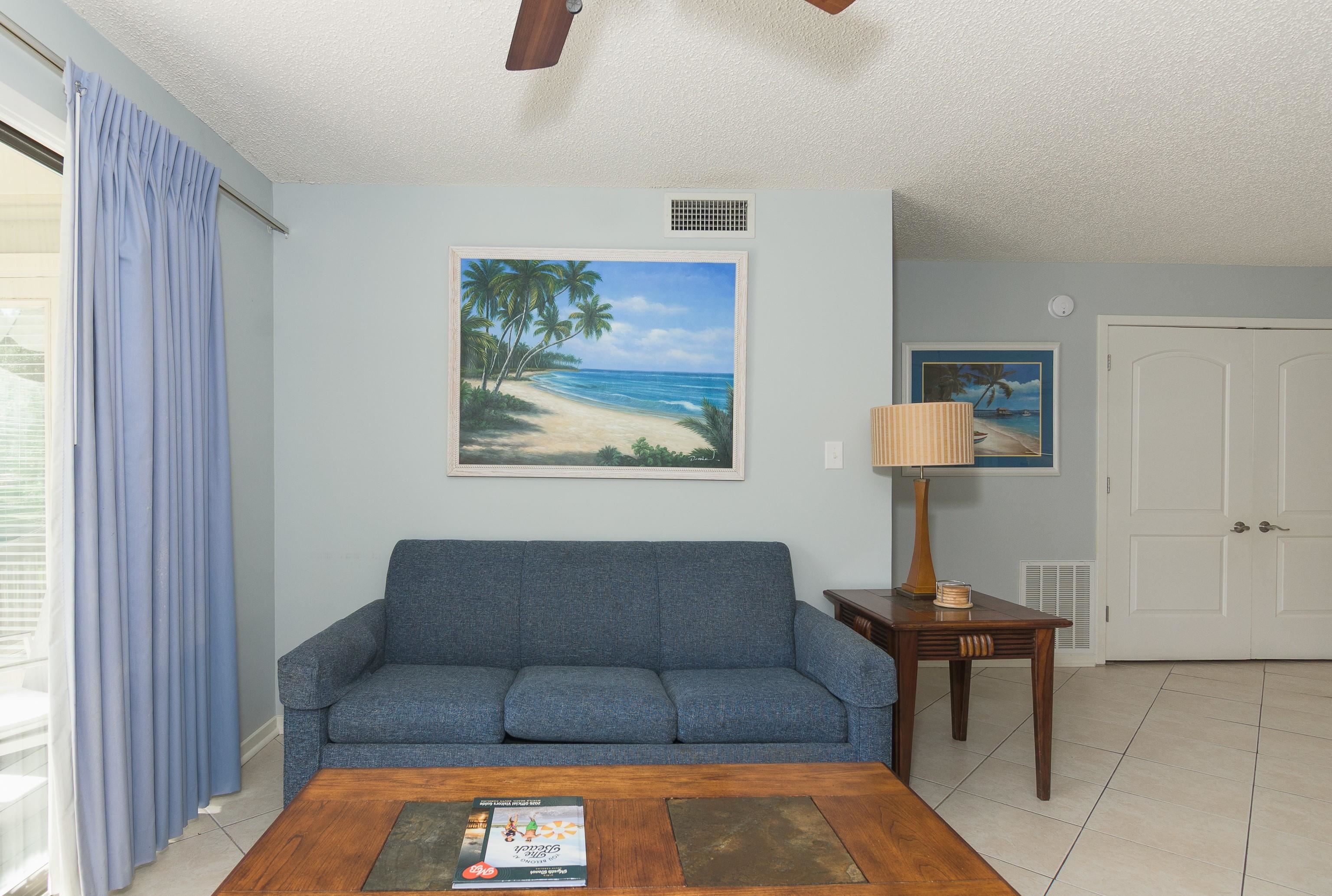 Living room with a blue sofa beneath a tropical beach painting and a wooden coffee table.