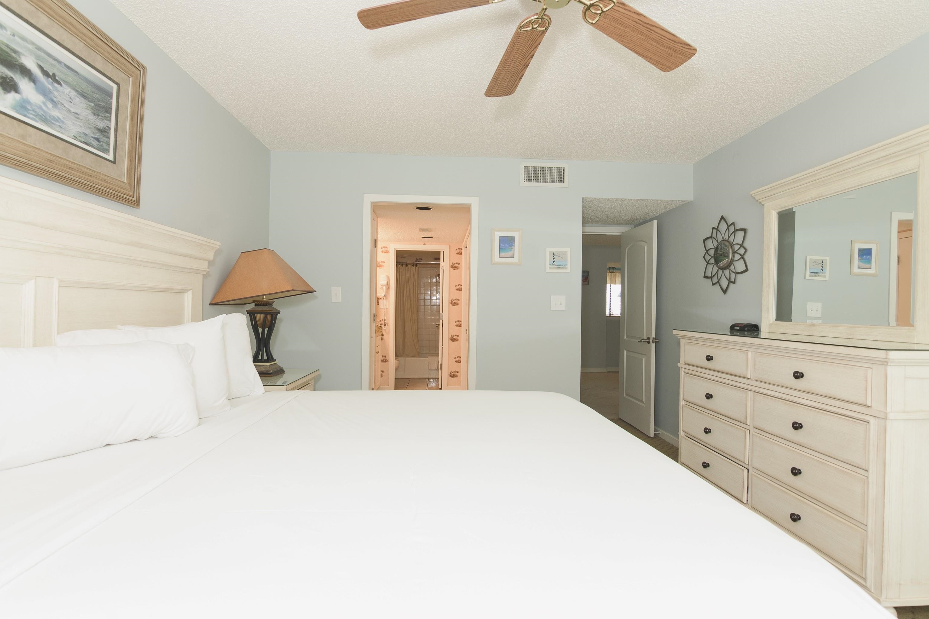 Bedroom with a white bed, pale wood dresser, ceiling fan, and ensuite bathroom doorway.