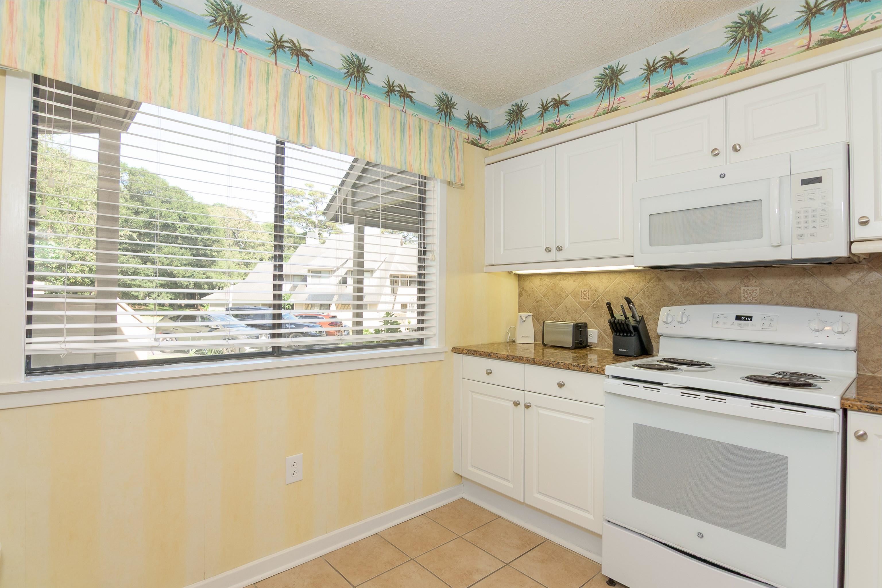 Kitchen with white cabinets and appliances, tan tile, and a large window beneath a palm-tree border.