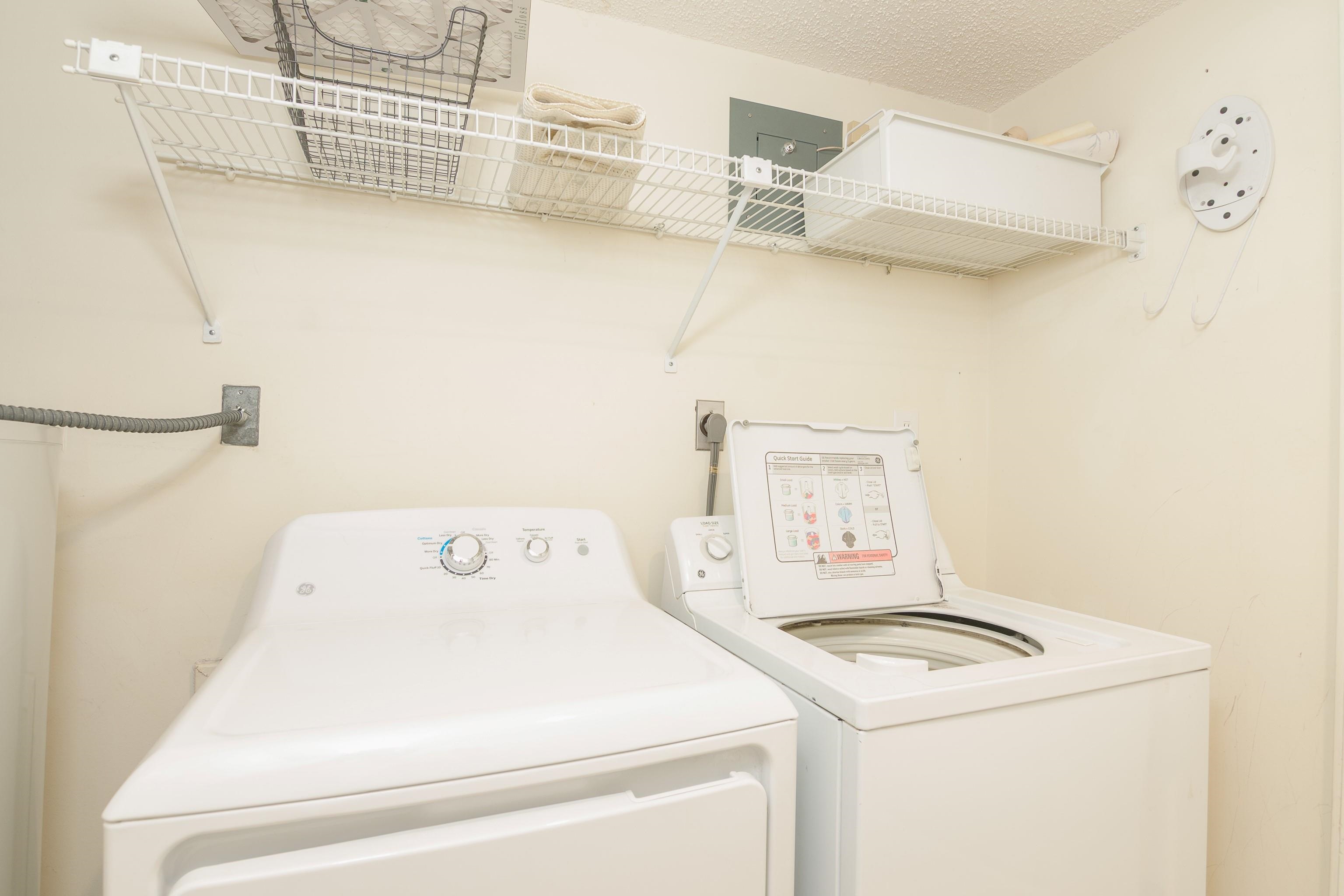 Laundry room with a top-loading washer and dryer beneath wire shelves.